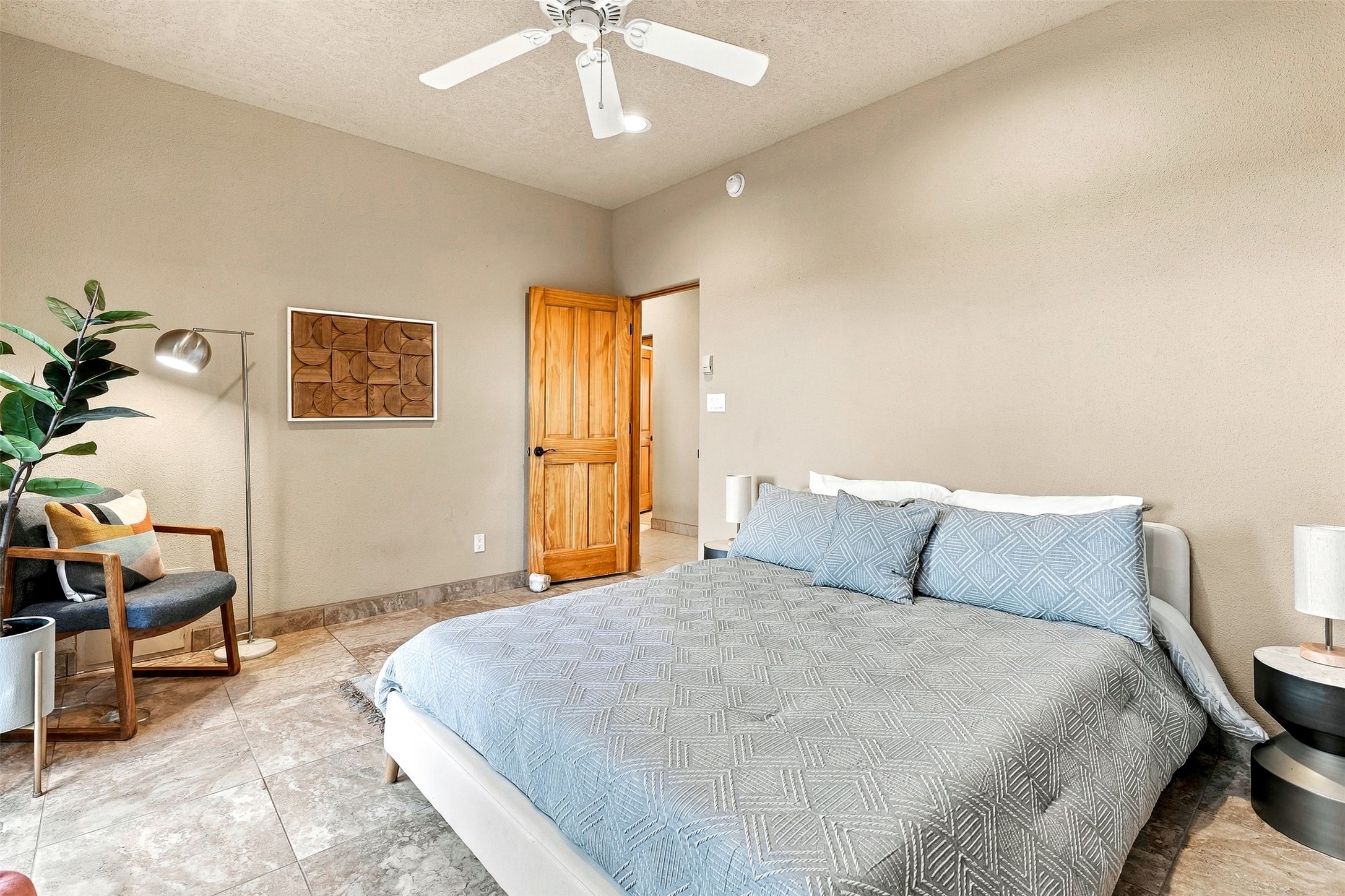 Staged bedroom with a gray-blue geometric quilt, wooden door, and stone tile floor.