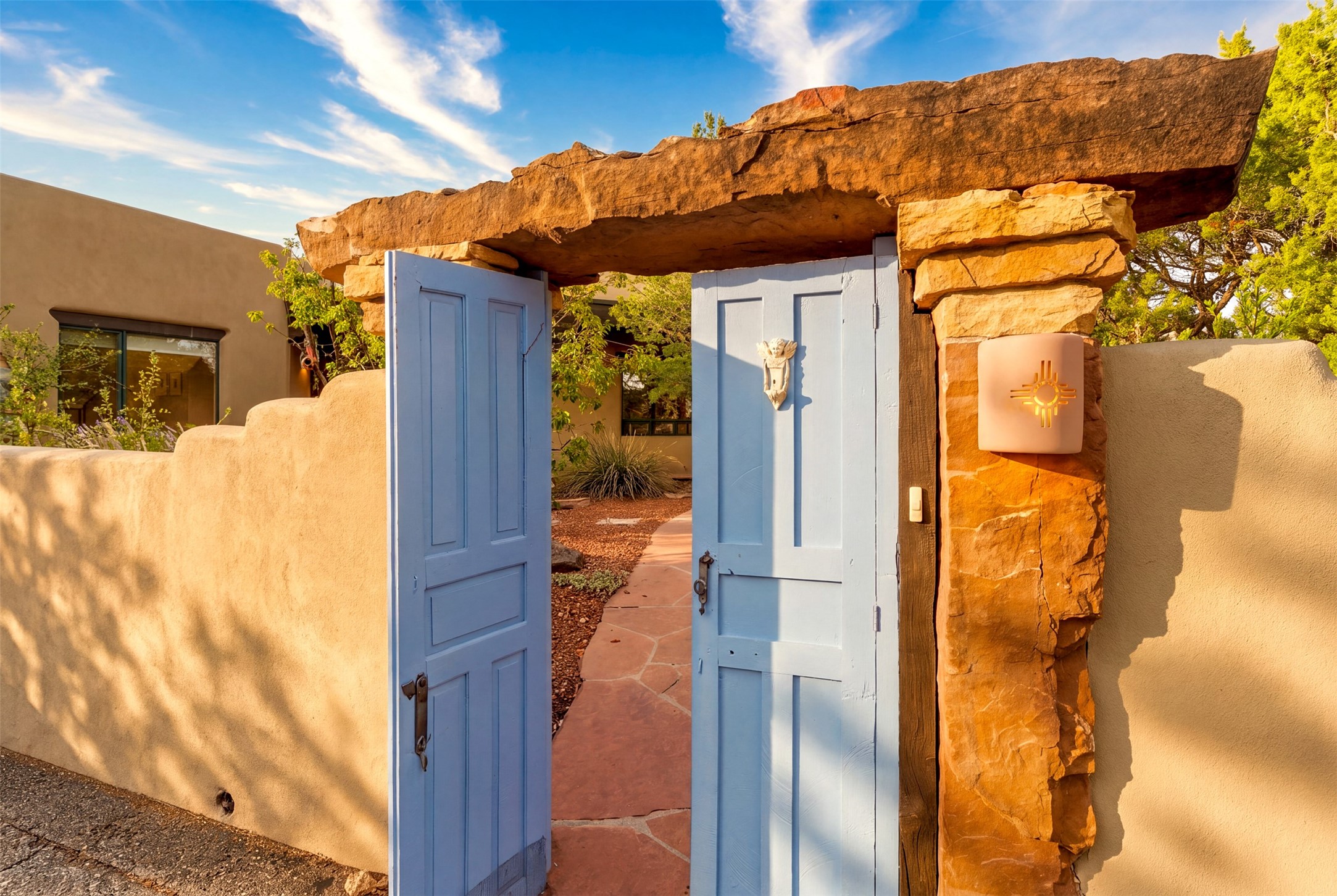 Adobe home entrance with open blue gates, a massive stone lintel, and a Zia sun emblem.