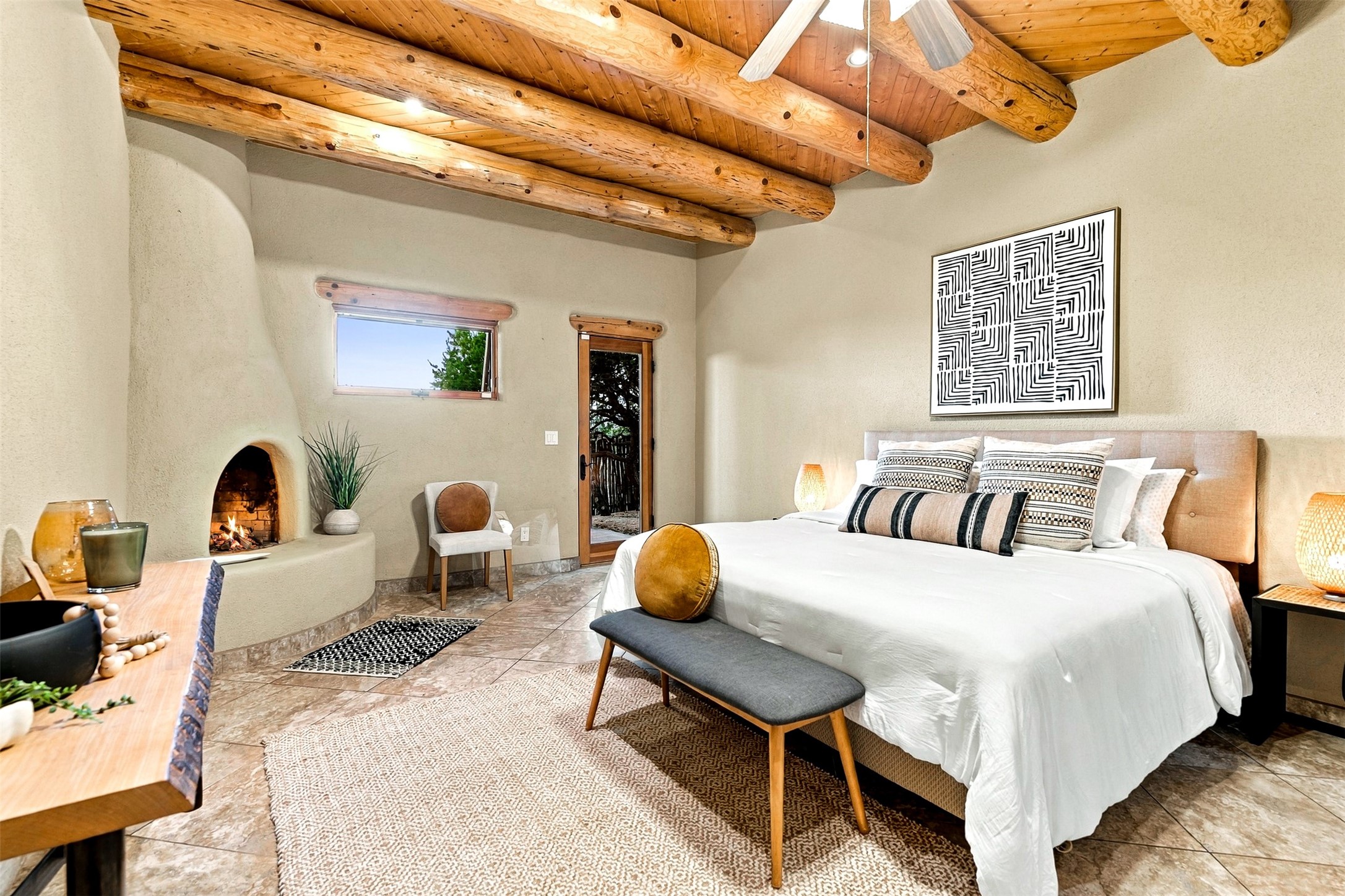 Bedroom with exposed timber beams, a kiva fireplace, and a king bed with a gray bench.