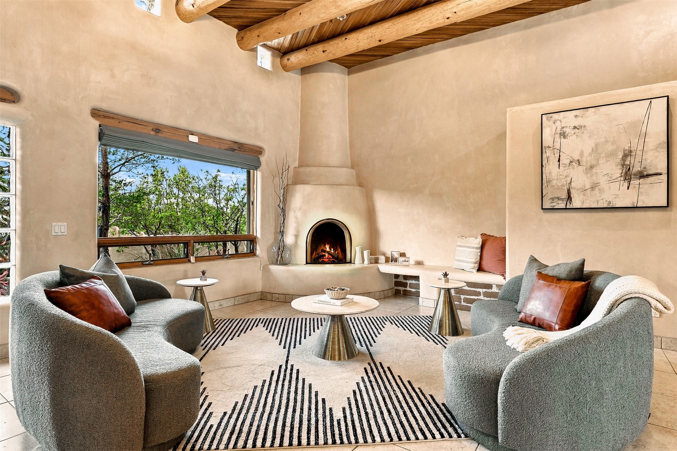 Living room with a lit kiva fireplace, exposed ceiling beams, and gray armchairs around a striped rug.