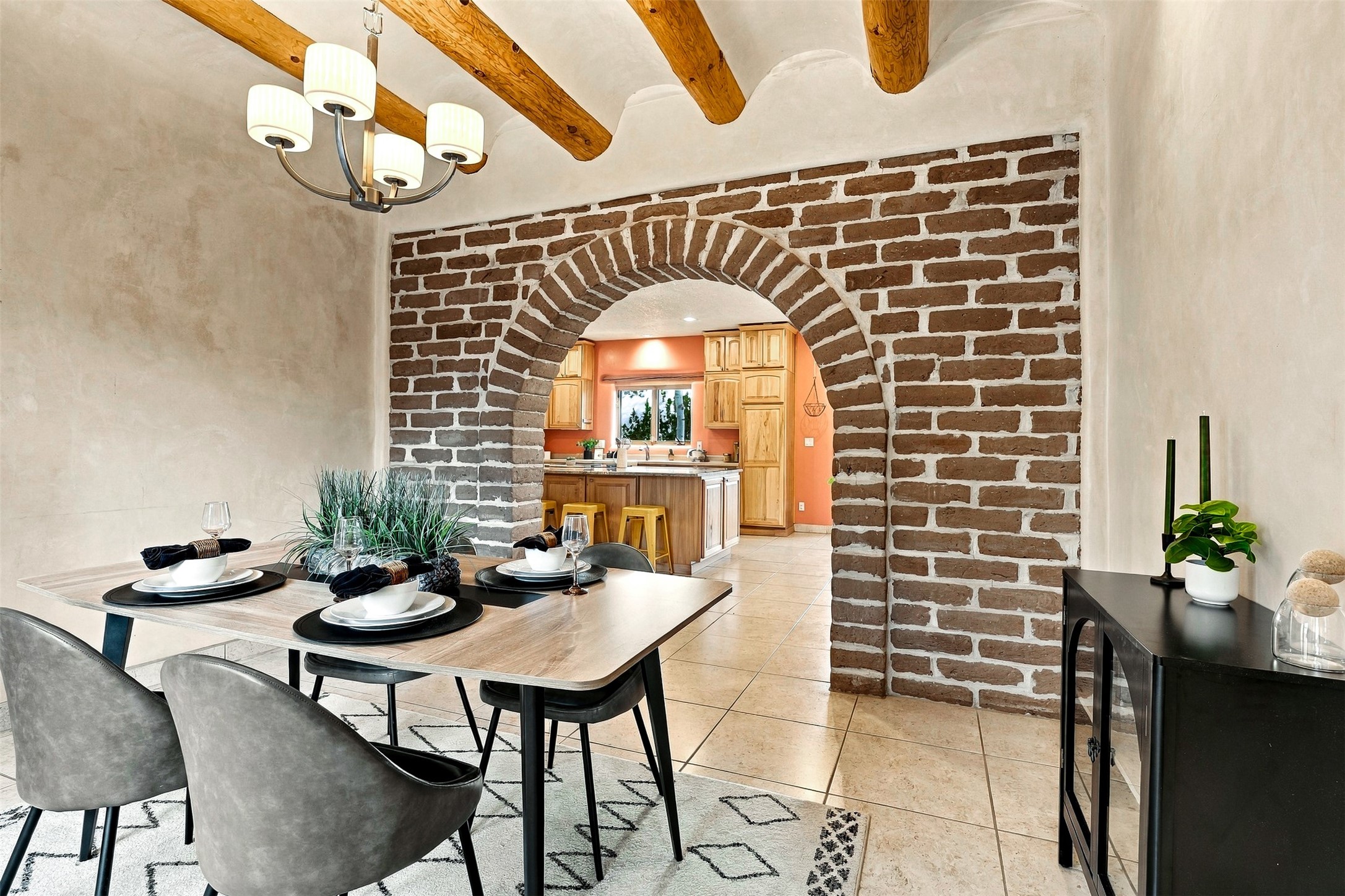 Dining room with a set table and exposed beams, looking through a brick arch into a wood-cabinet kitchen.