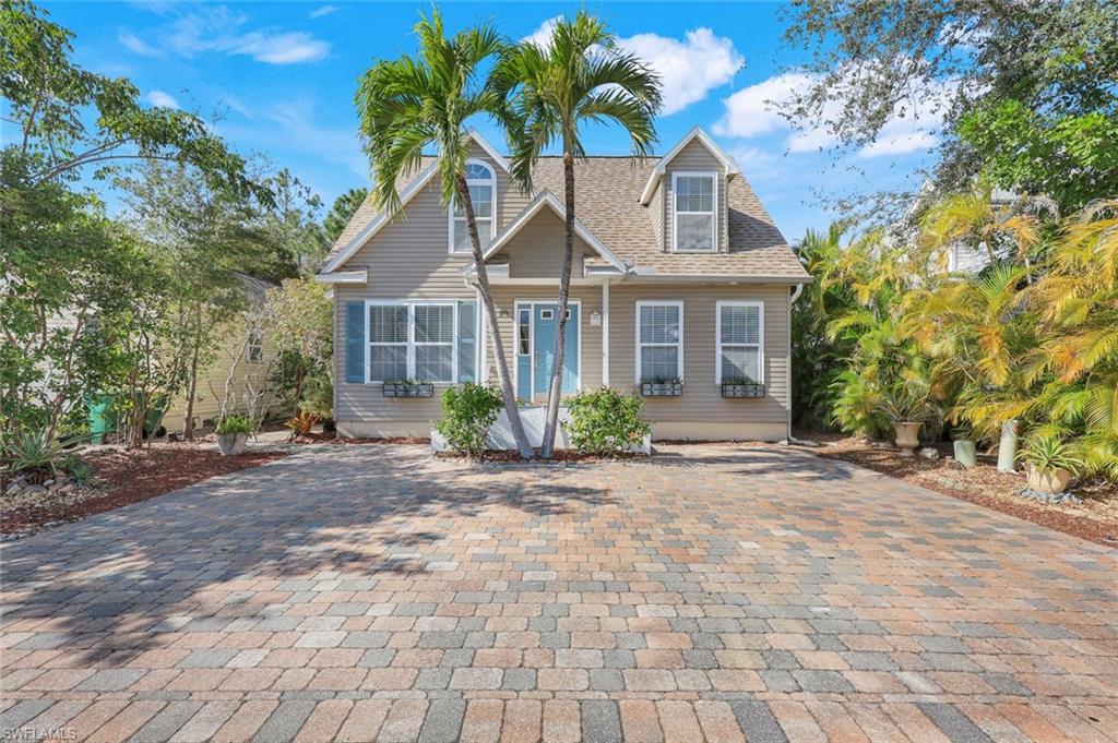 Gray-sided home with a blue entry door, dormer windows, and a wide paver driveway framed by palm trees.