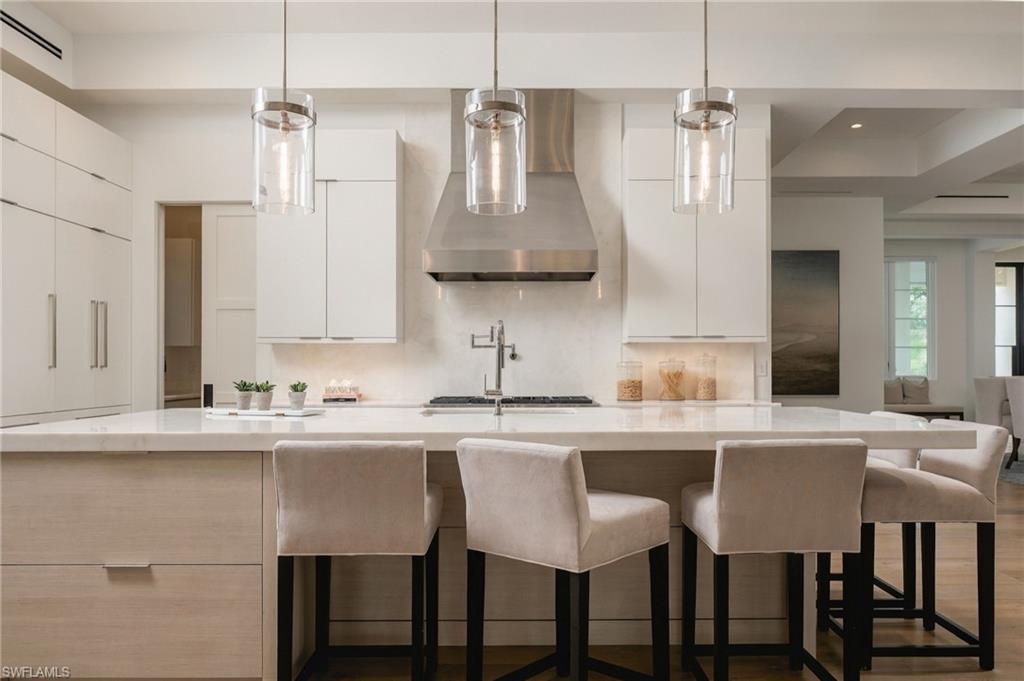 Modern white kitchen with a large island, upholstered bar stools, and glass pendant lights.