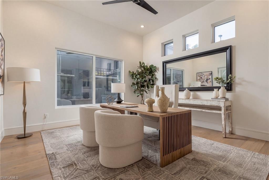 Staged home office with a wood desk, upholstered chairs, large mirror, and high windows.
