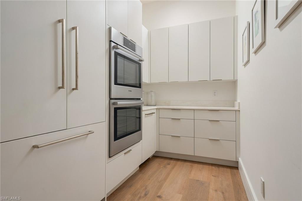 Modern white kitchen with stainless steel double ovens, sleek cabinetry, and wood flooring.