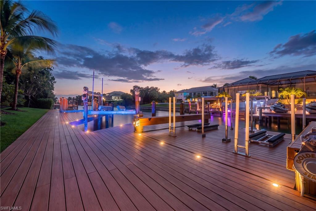 Waterfront dock and broad wooden deck illuminated at dusk, with boats and homes across the water.