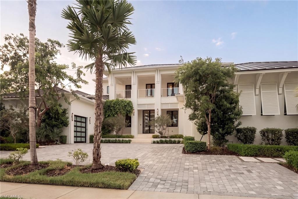 Modern two-story white home with a wide paver driveway, balconies, and tropical landscaping.