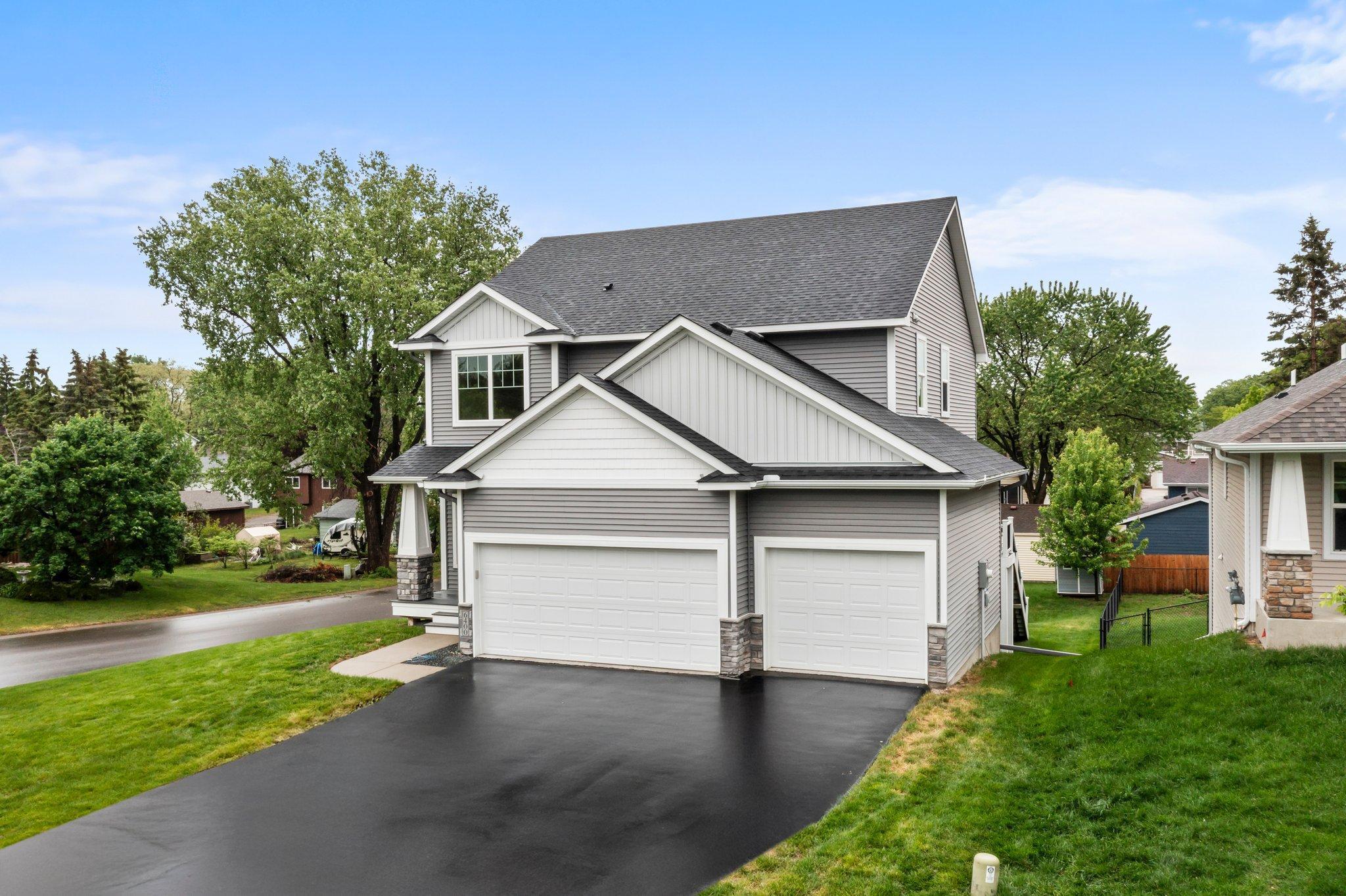 Two-story gray house with white gables, stone accents, and a three-car garage beside a freshly paved driveway.