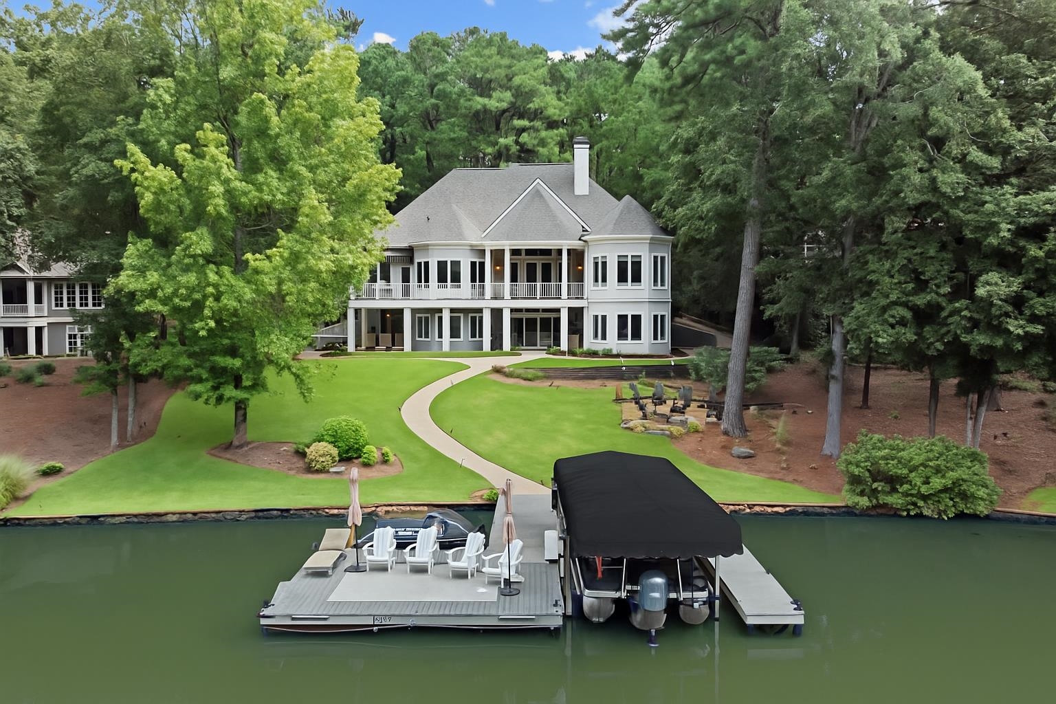 Lakefront house with a landscaped lawn, dock, covered boat, and wooded surroundings.