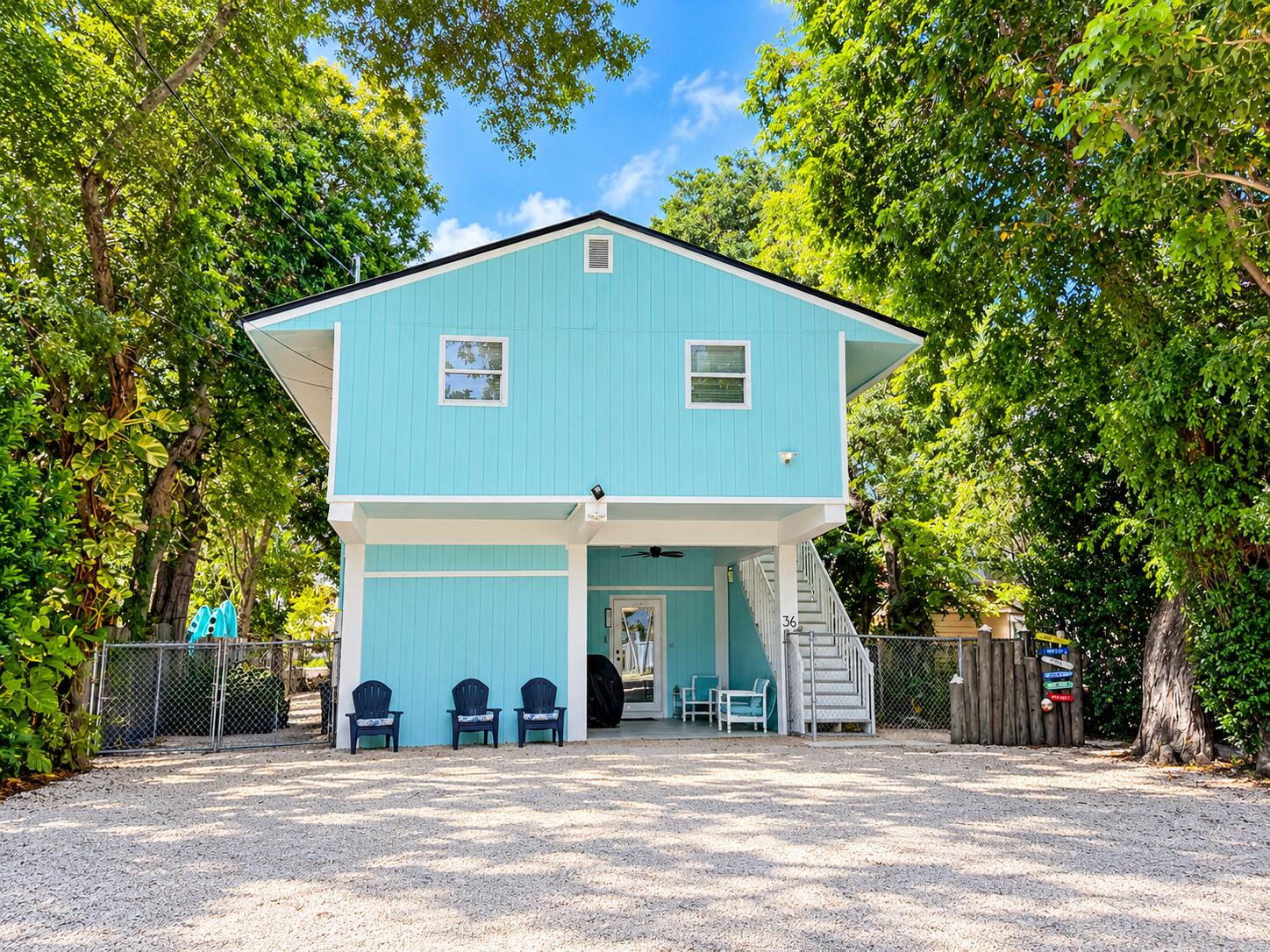 Turquoise raised house with white exterior stairs and a gravel driveway beneath tropical trees.