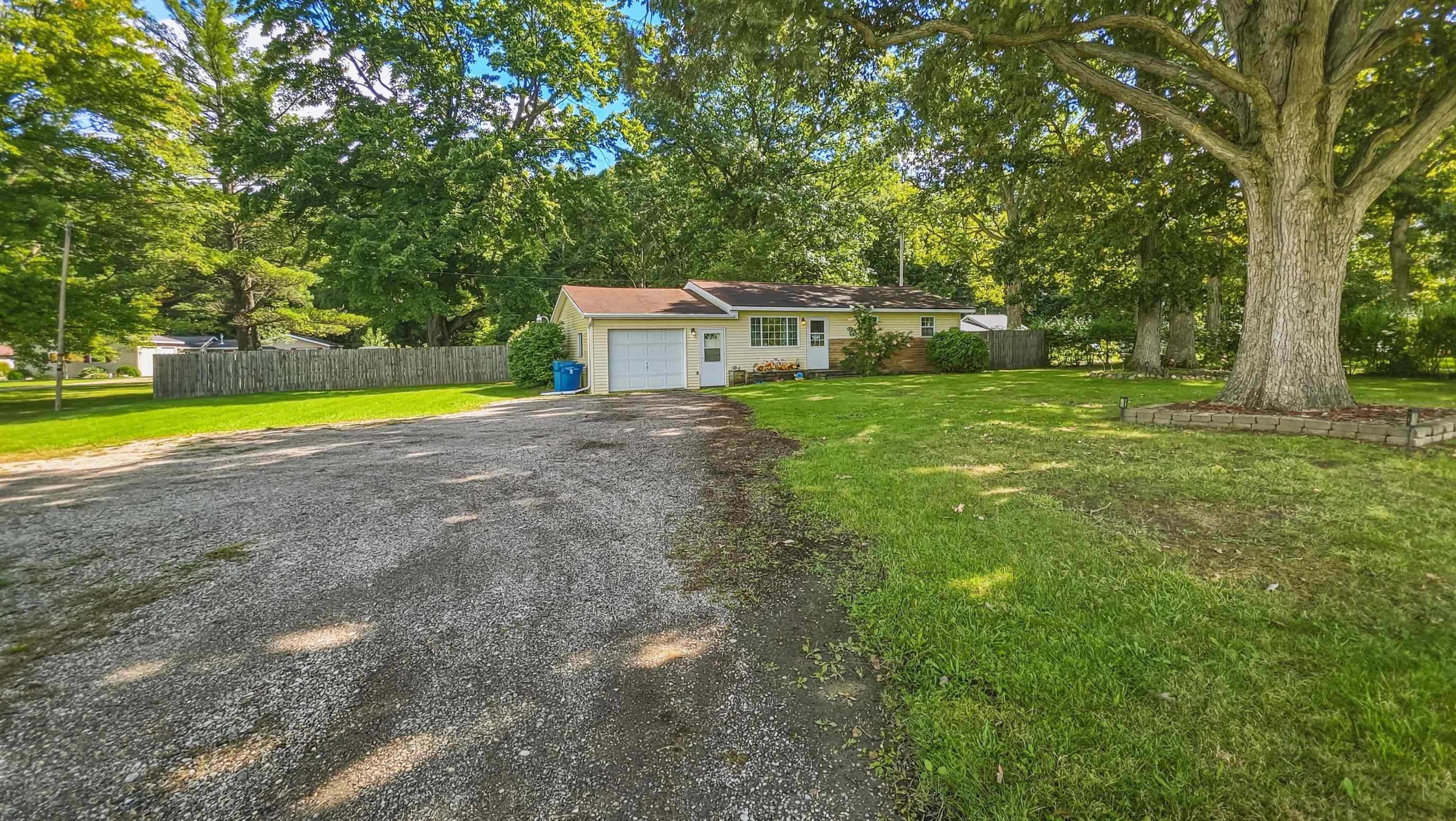 Single-story yellow home with attached garage, gravel driveway, and large shade trees.