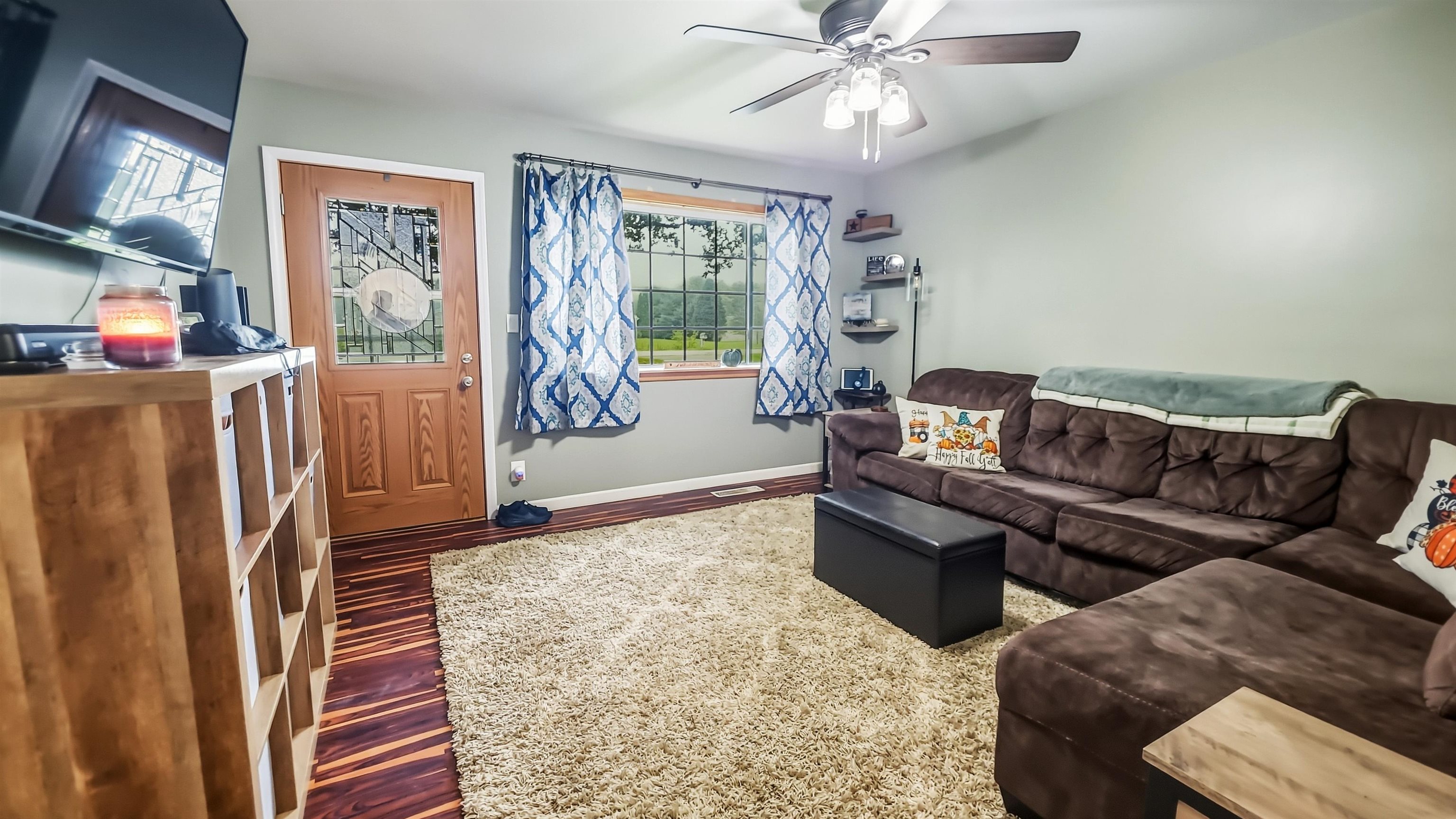 Living room with a brown sectional, cream shag rug, striped hardwood floor, and blue patterned curtains.