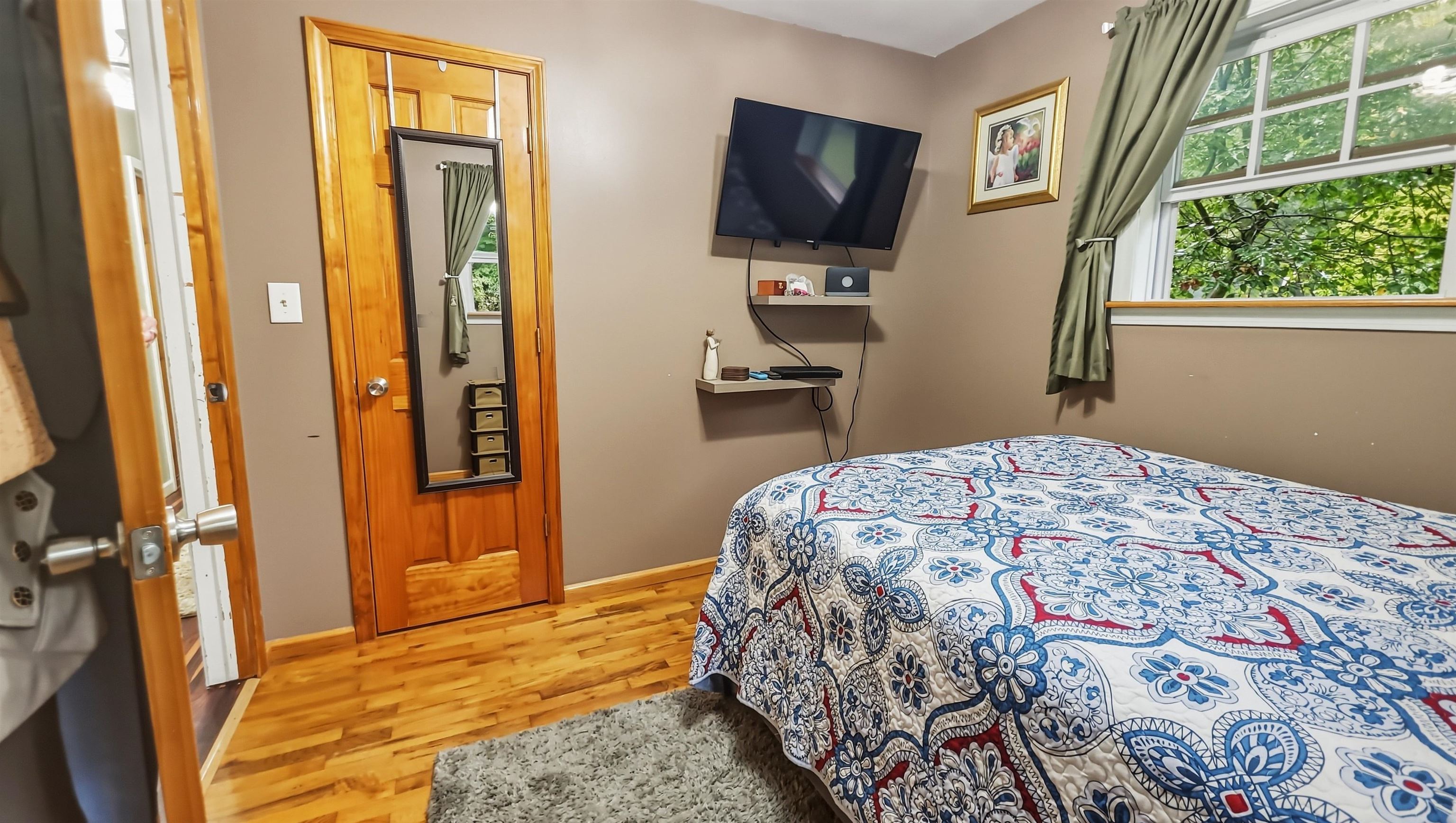 Bedroom with a colorful patterned bedspread, hardwood floors, and a large curtained window.