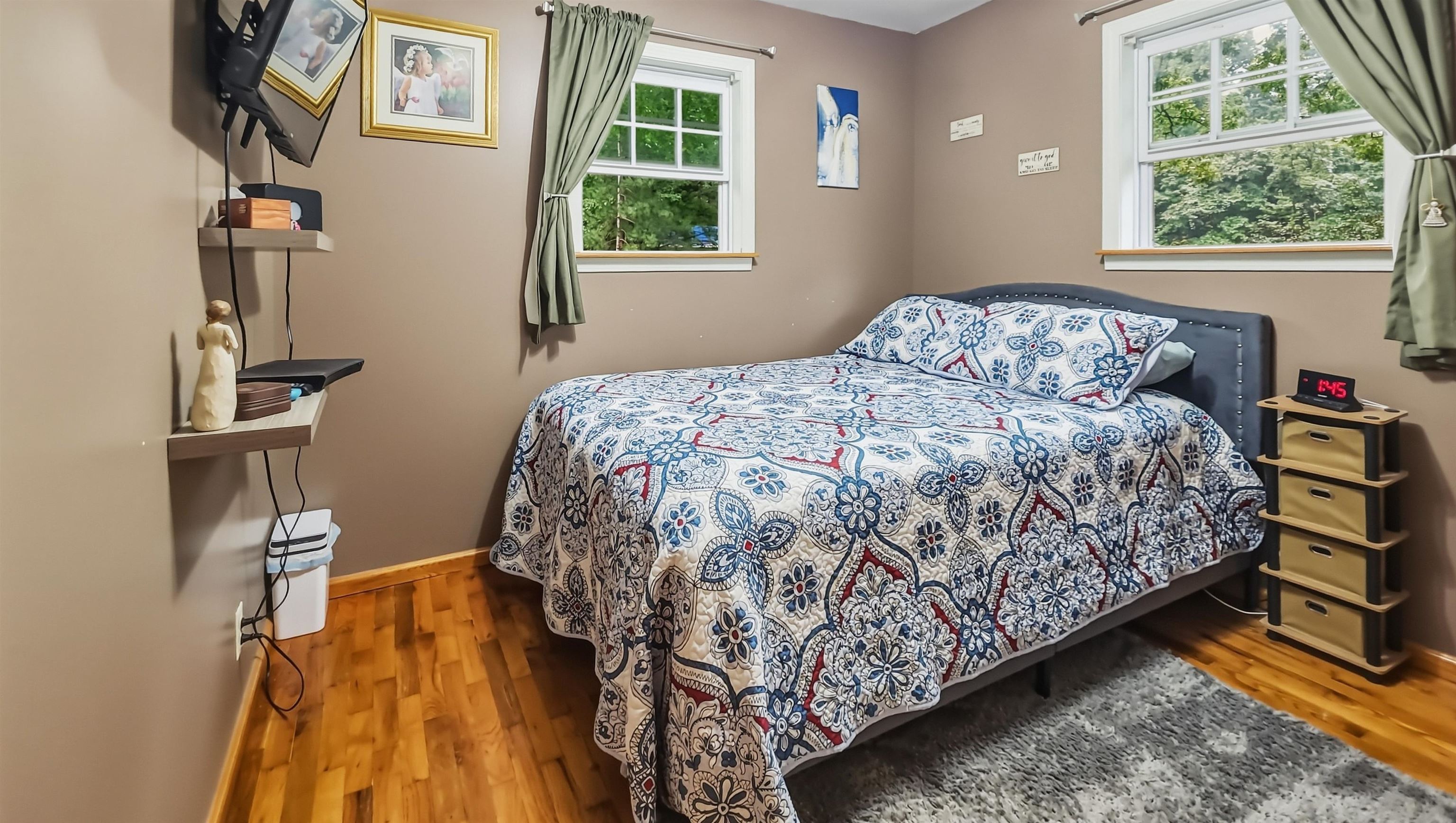 Bedroom with a patterned bedspread, two curtained windows, hardwood floors, and a gray rug.
