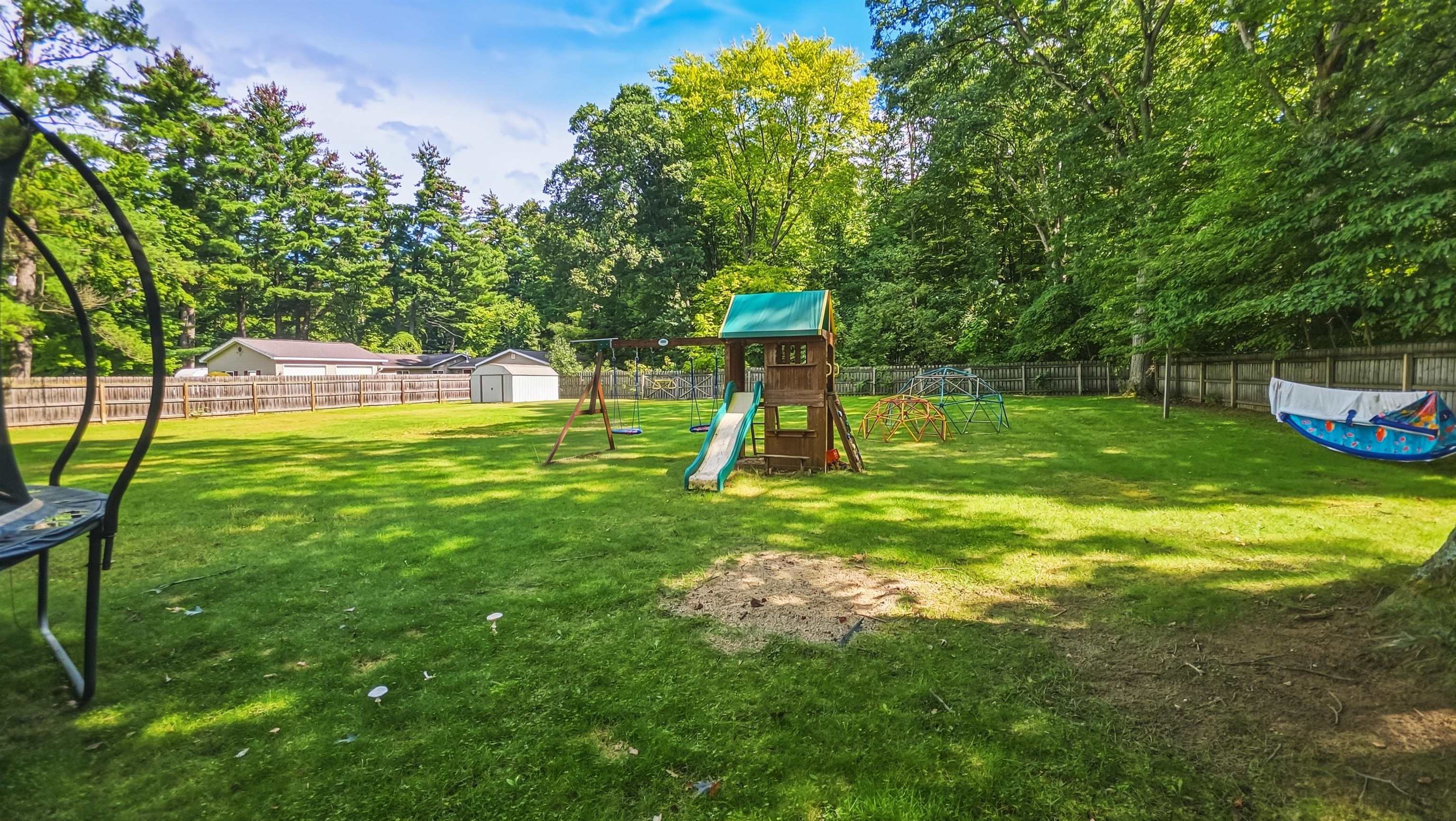 Fenced backyard with a wooden playset, swings, climbing domes, and a broad lawn.