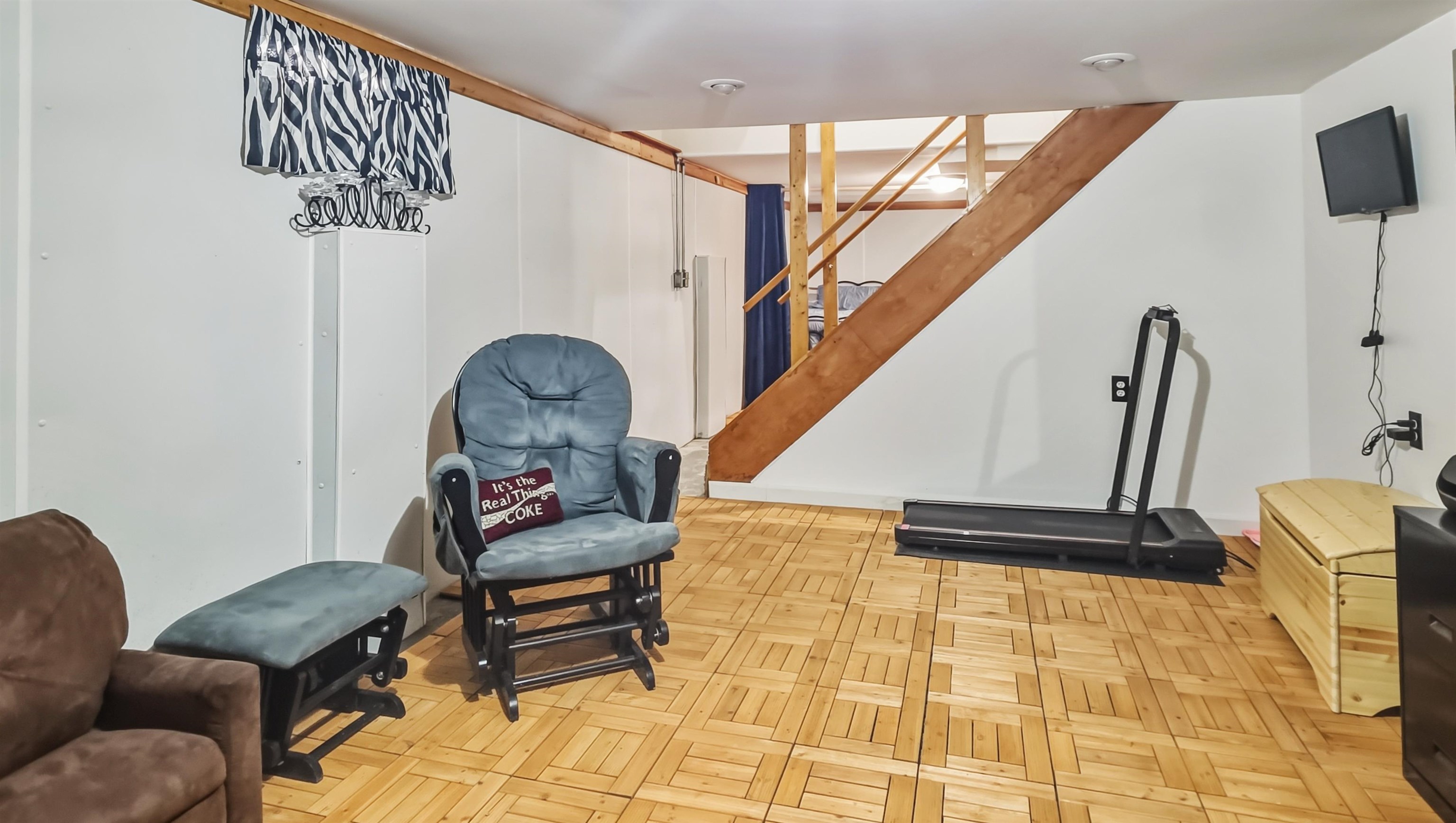 Recreation room with a wooden staircase, treadmill, rocking chairs, and patterned wood flooring.