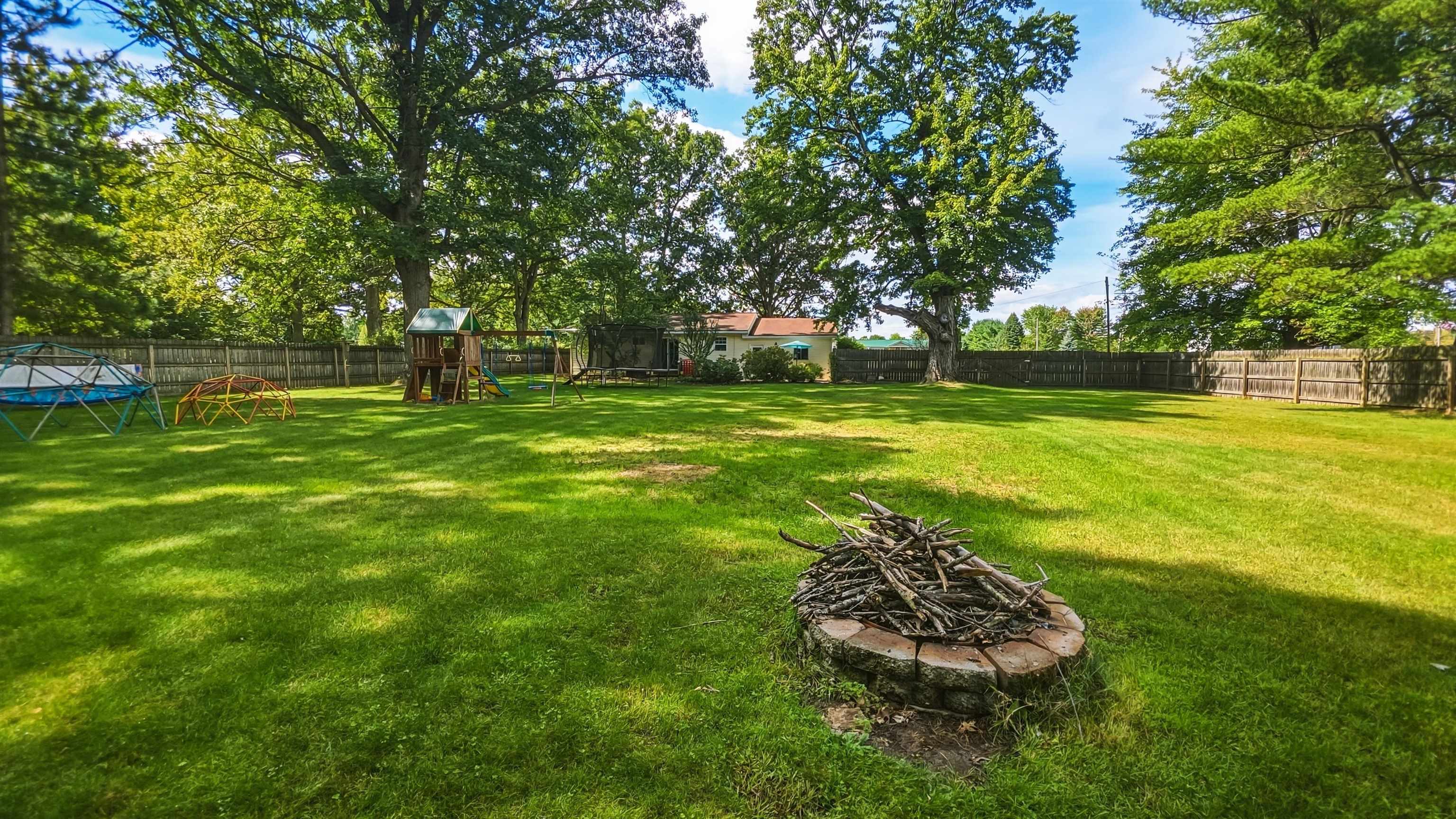 Spacious fenced backyard with mature trees, a stone fire pit, and children's play equipment.