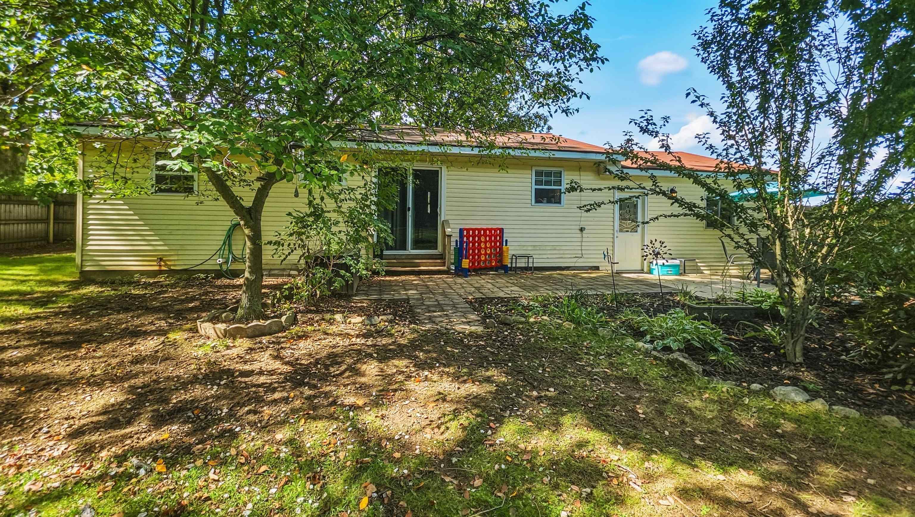 Rear of a yellow-sided house with a paved patio, outdoor chairs, and leafy backyard trees.