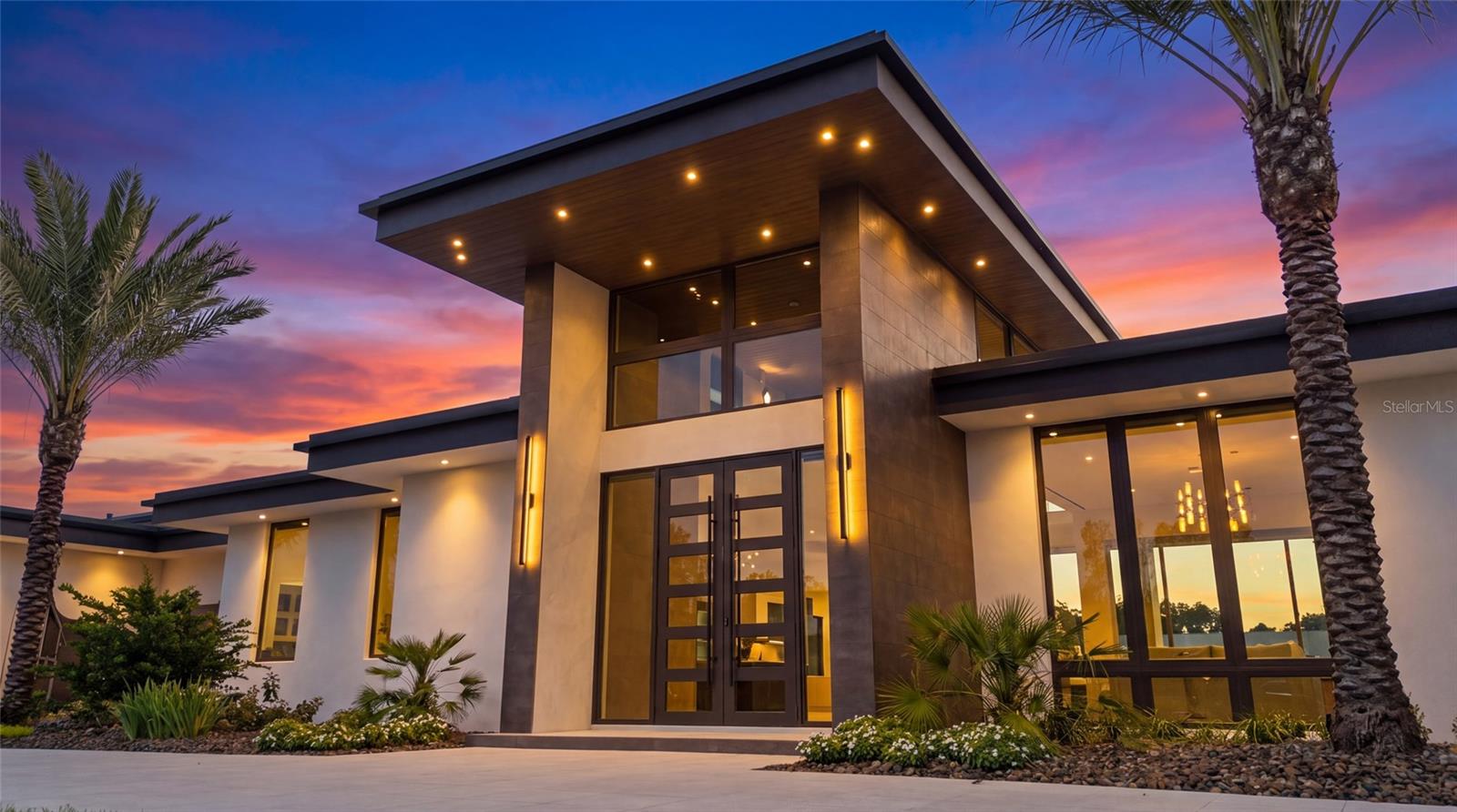 Modern home with an angular roof and glass walls, lit at dusk among palm trees.