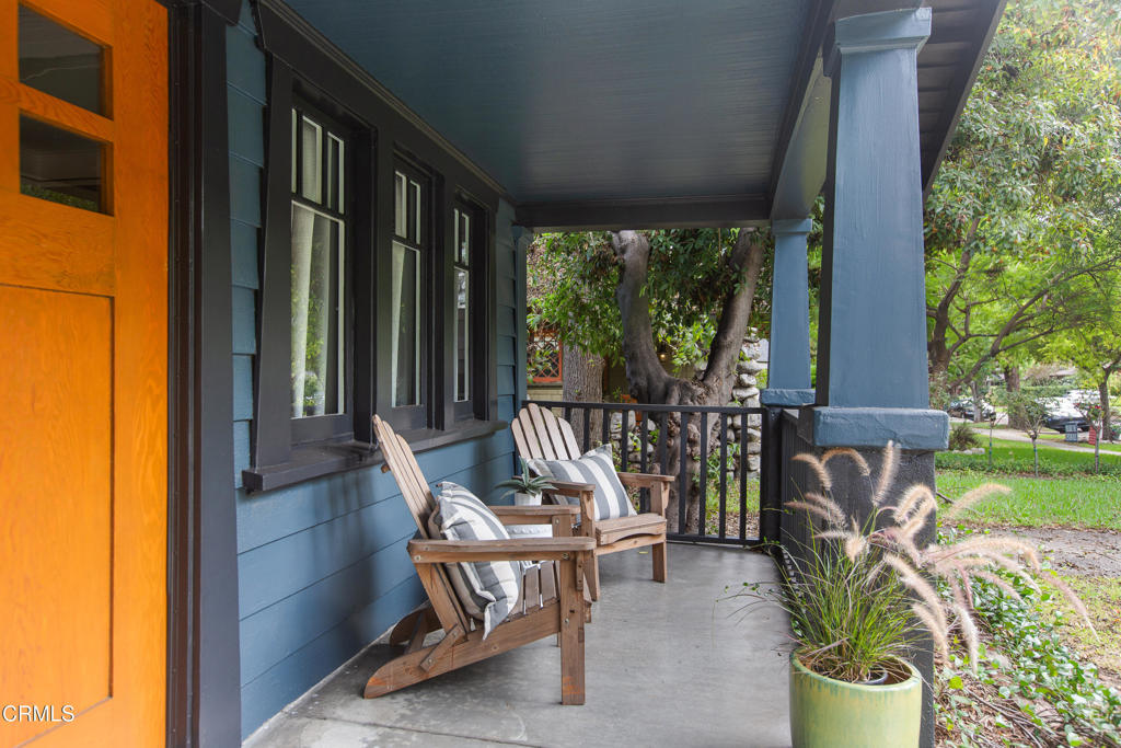 Covered porch with blue siding, an orange door, and two wooden chairs beside a potted grass.