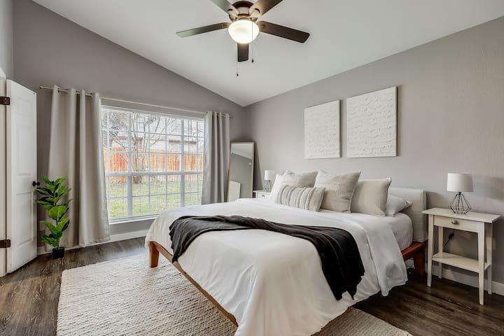 Staged bedroom with a white bed, dark throw, vaulted ceiling, and large curtained window.