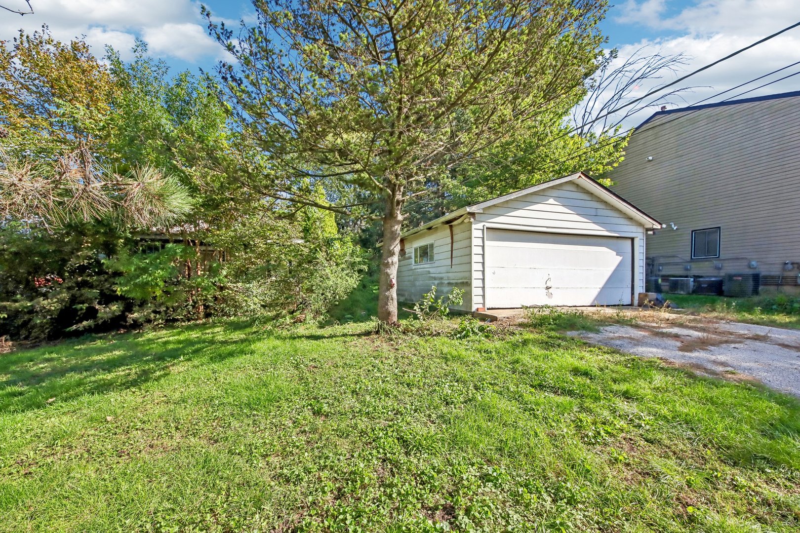 Detached white garage beside a larger home, with a mature tree and grassy yard.