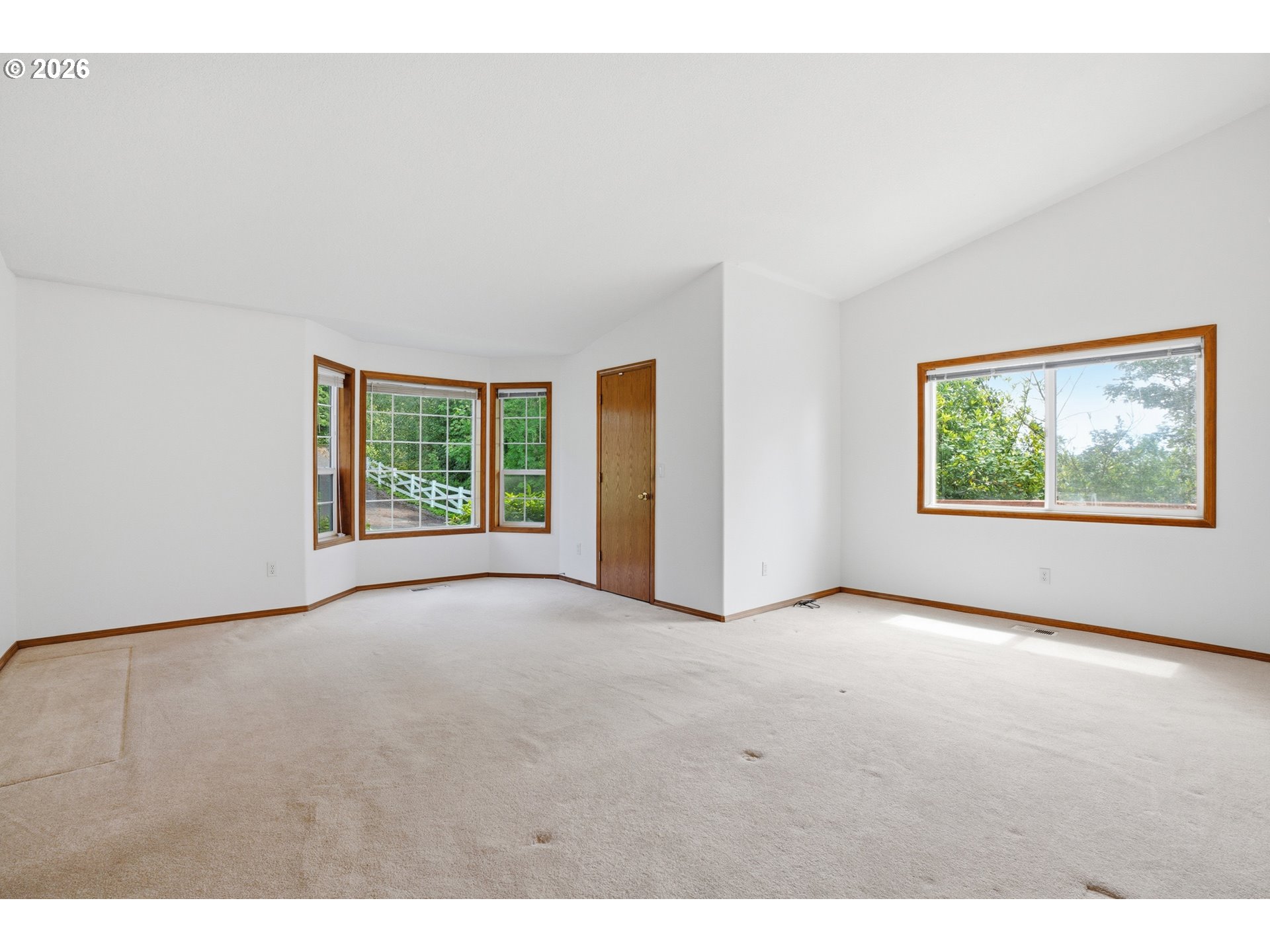Empty carpeted room with wood-trimmed windows overlooking greenery and a white fence.
