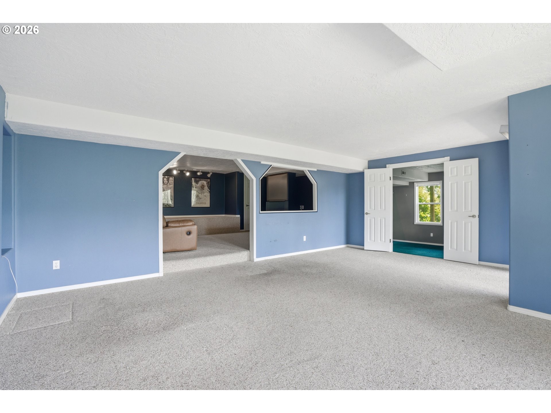 Spacious carpeted room with blue walls, archways to a sitting area, and open double doors to another room.