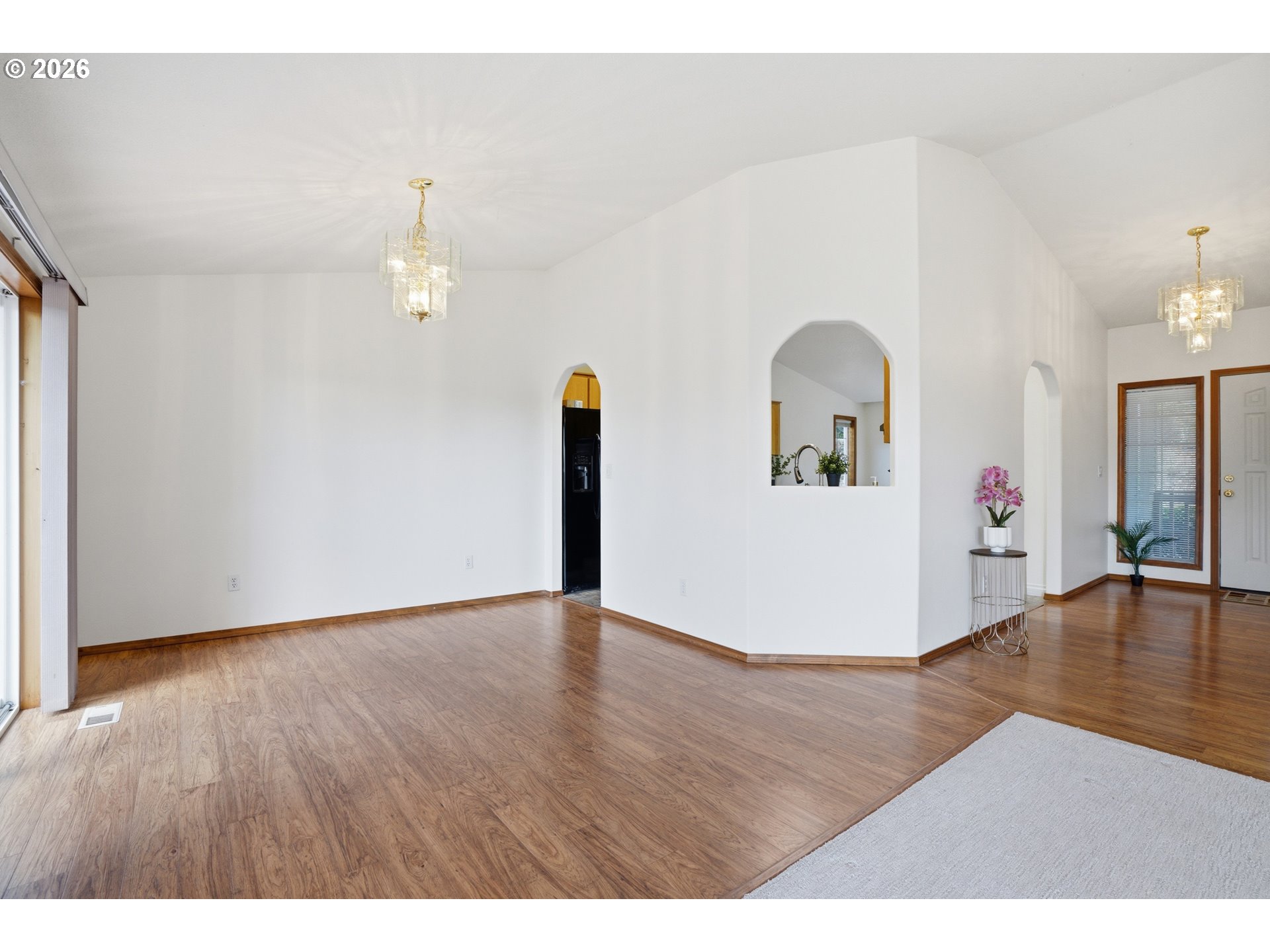 Mostly empty living and dining area with wood floors, arched openings, and two chandeliers.