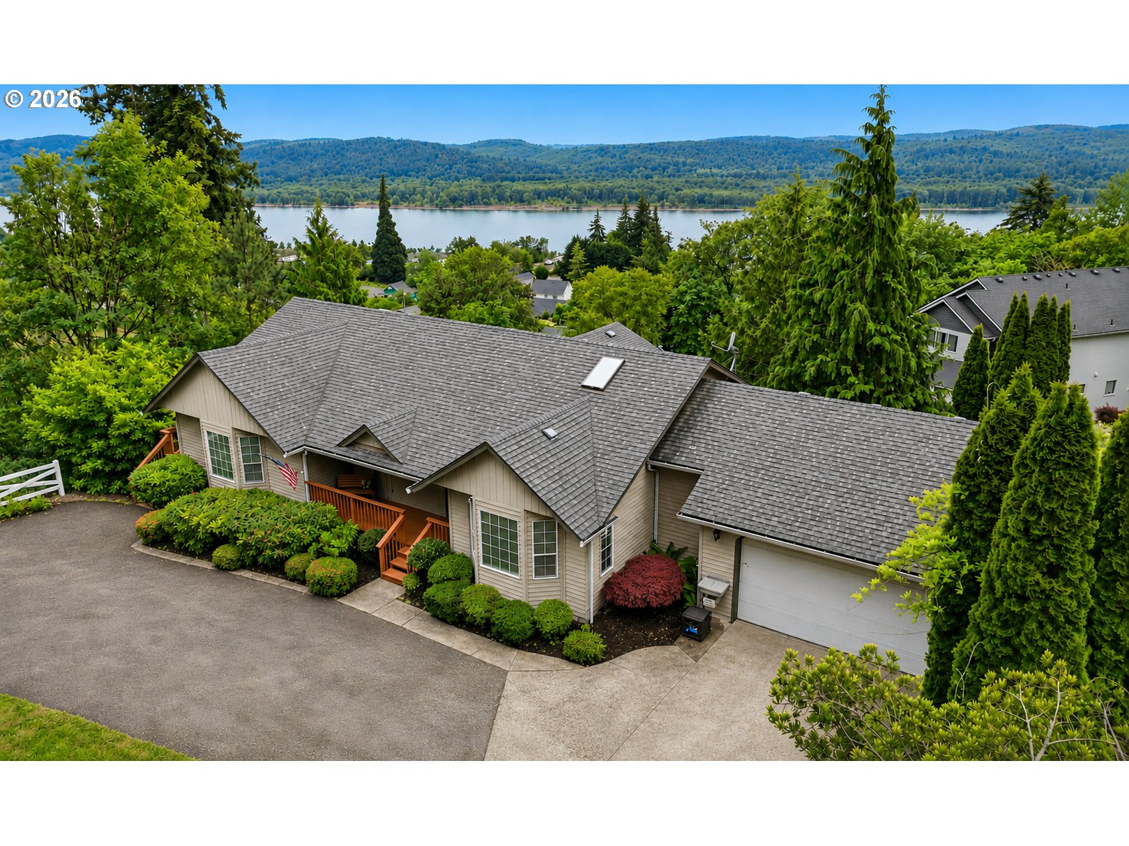 Beige home with a gray roof and landscaped grounds, overlooking a lake and forested hills.