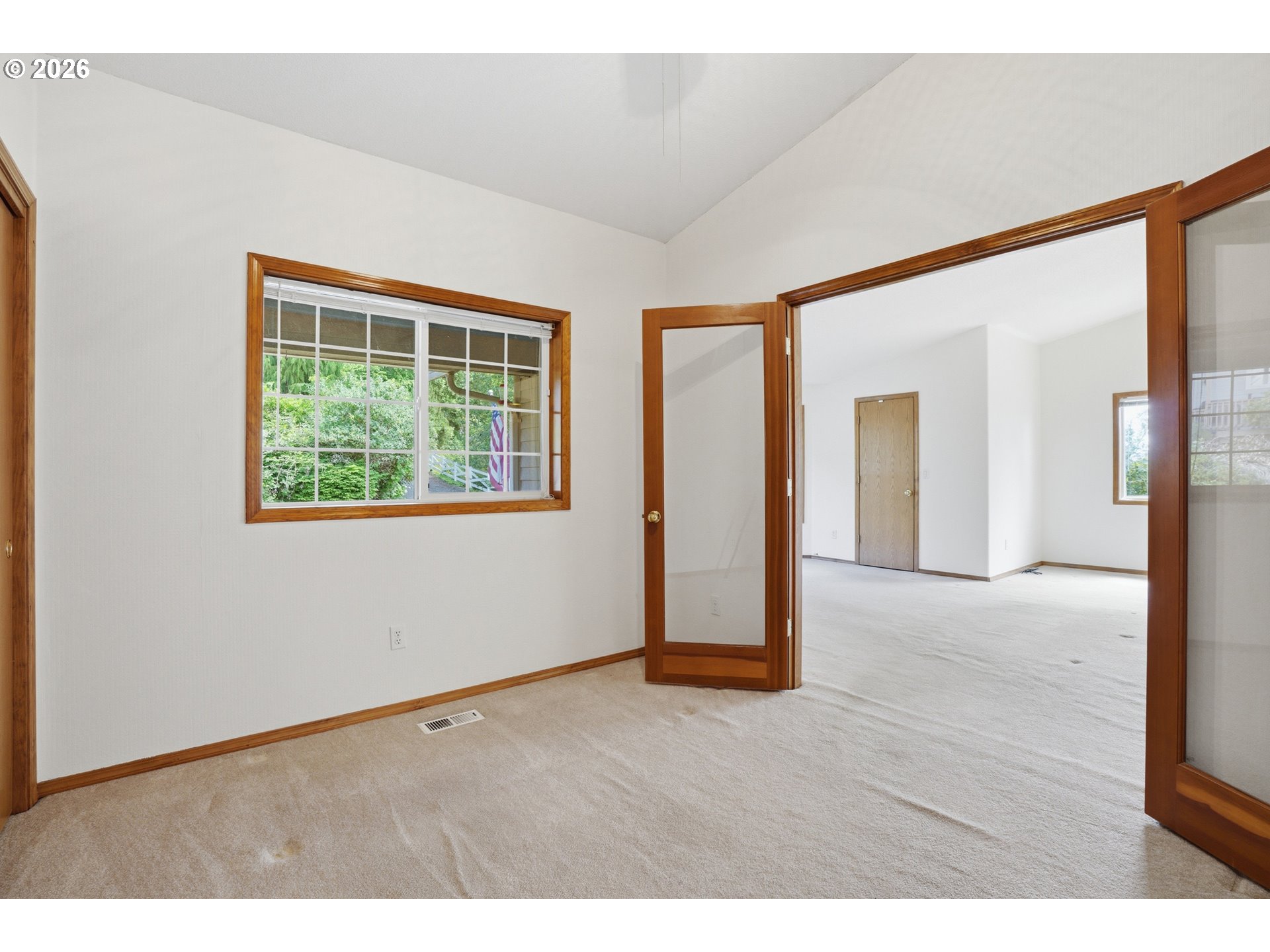 Empty carpeted room with wood-trimmed windows and an open doorway to a larger room.