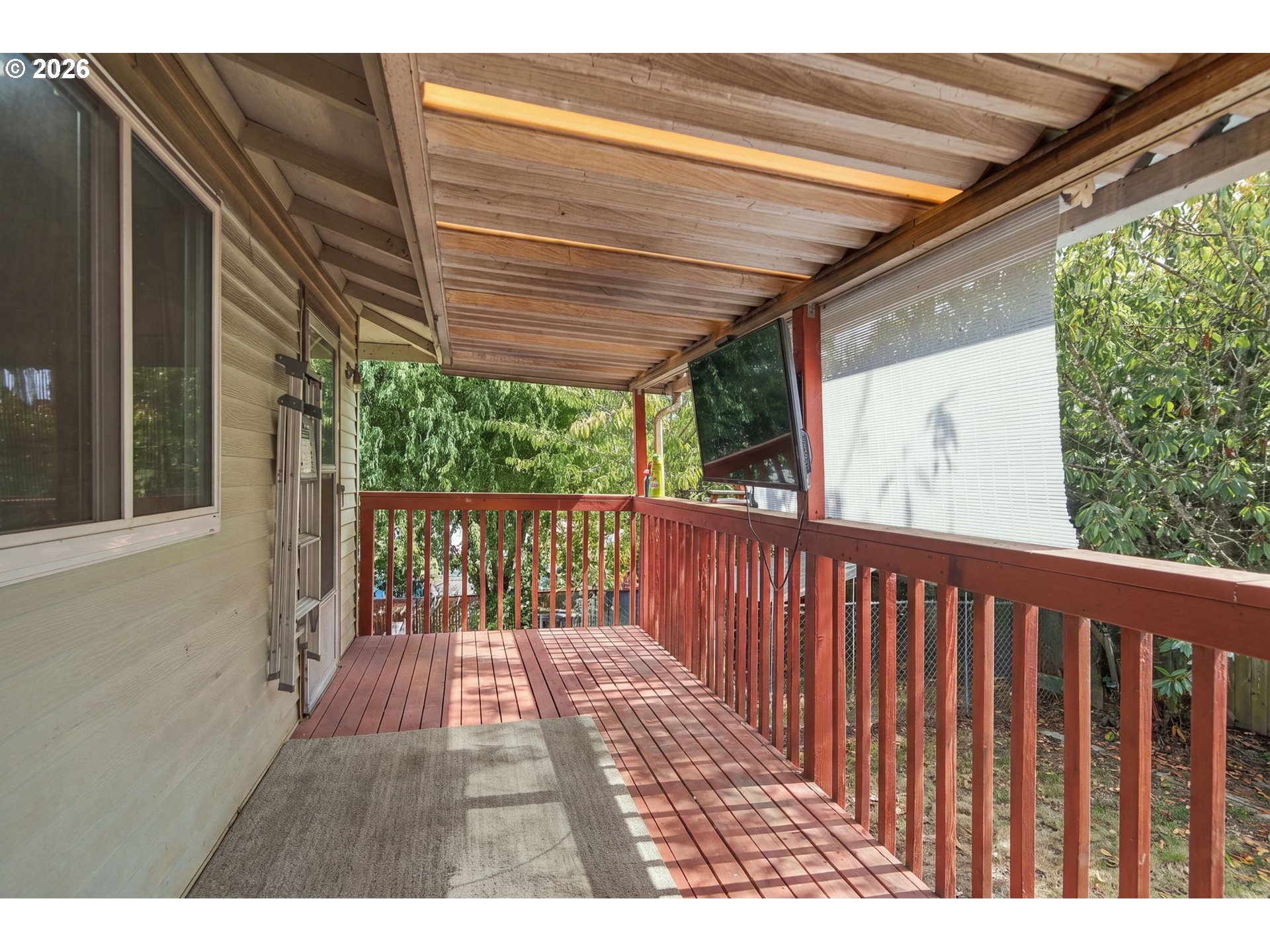 Covered wooden deck with red railings, overhead roof, and mounted television overlooking trees.