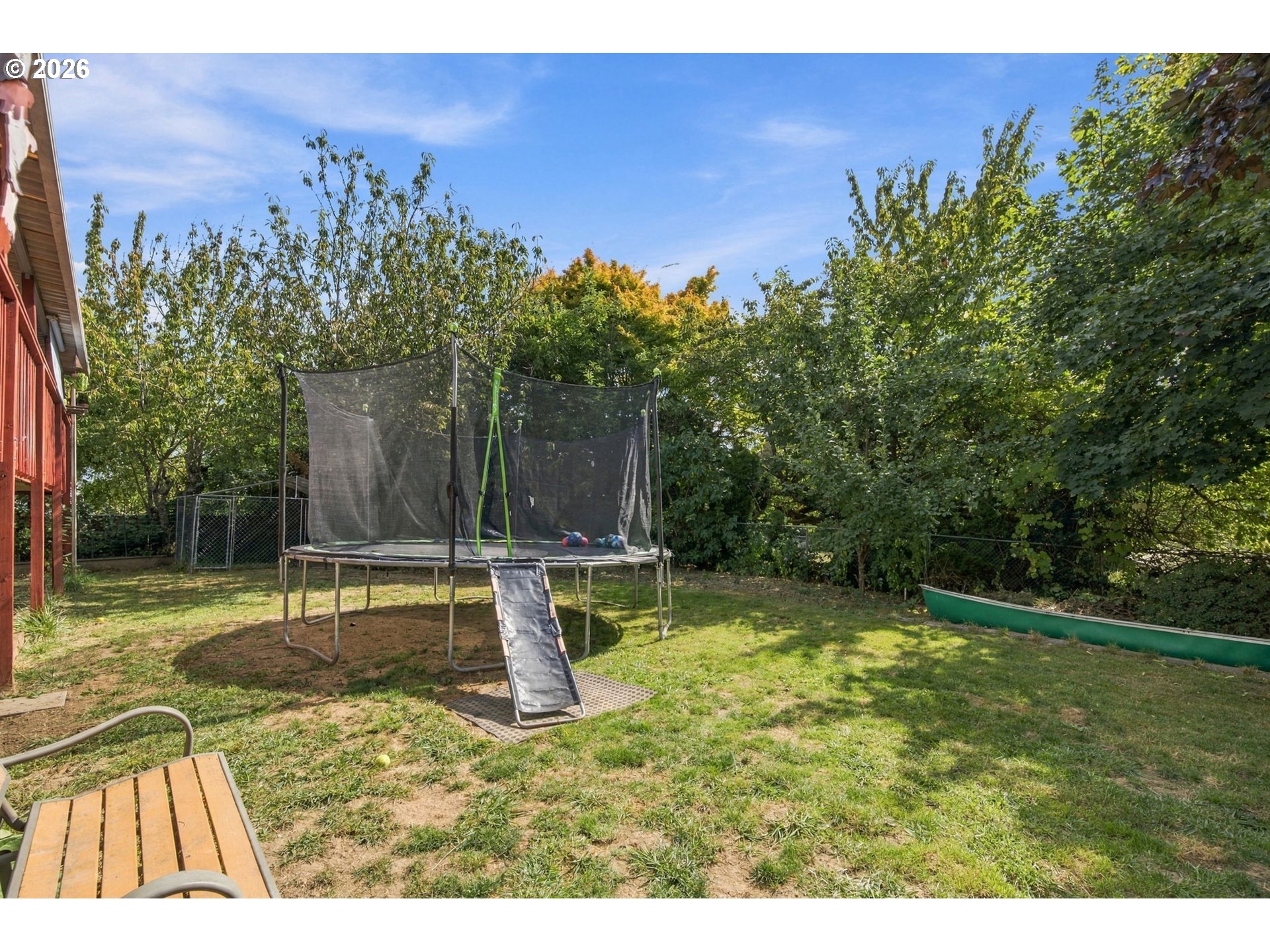Fenced backyard with a large netted trampoline and ramp beneath mature trees.