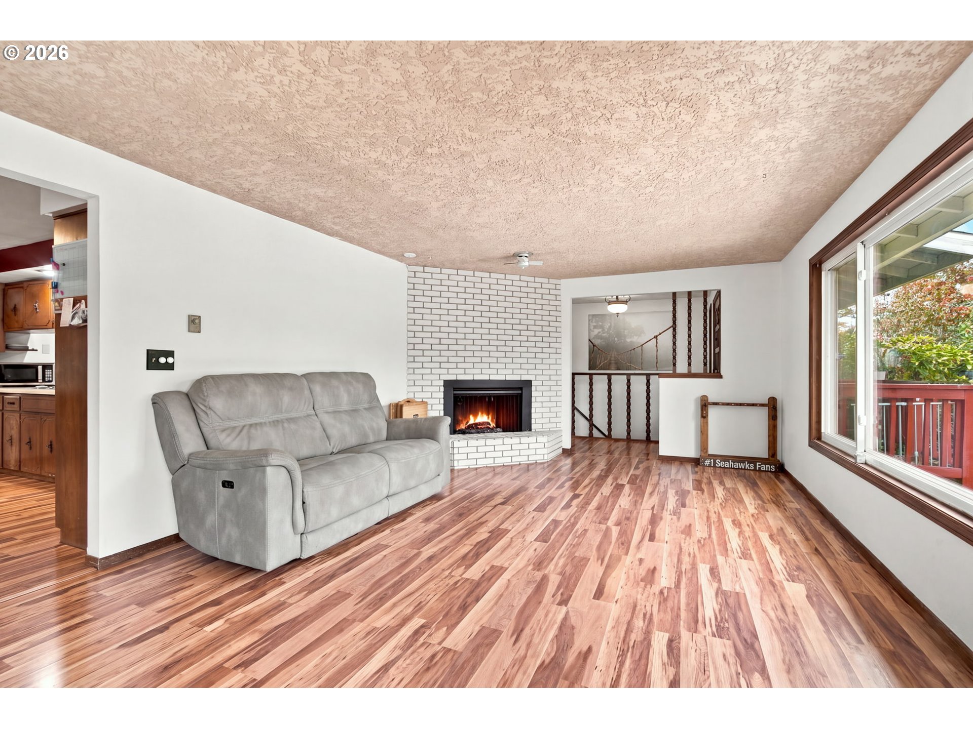 Living room with gray reclining sofa, white brick fireplace, wood-look floors, and a large window overlooking a deck.