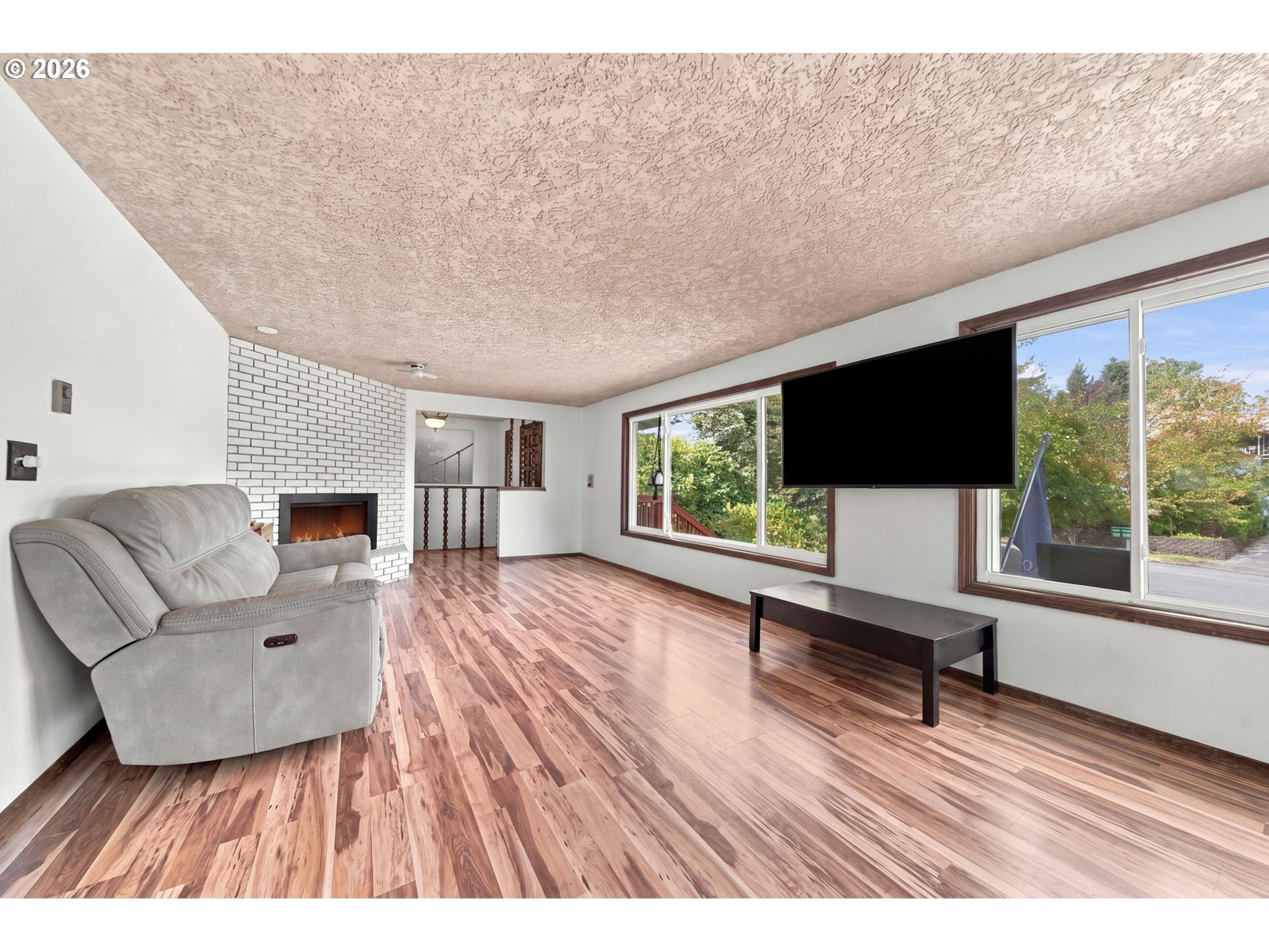 Living room with a white brick fireplace, gray recliner, and large windows overlooking greenery.
