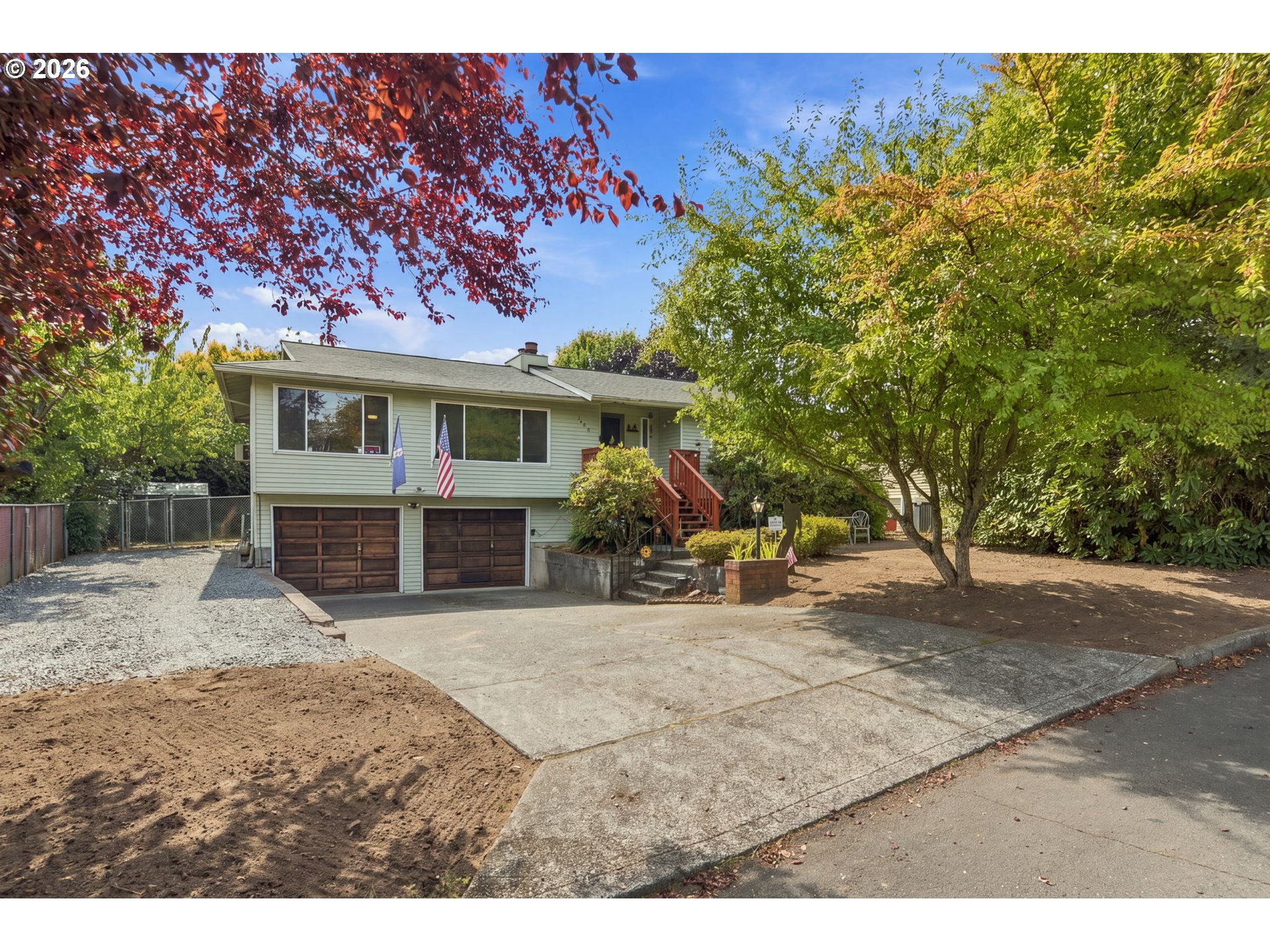 Light green split-level house with two garage doors, a red entry staircase, and mature trees framing the driveway.