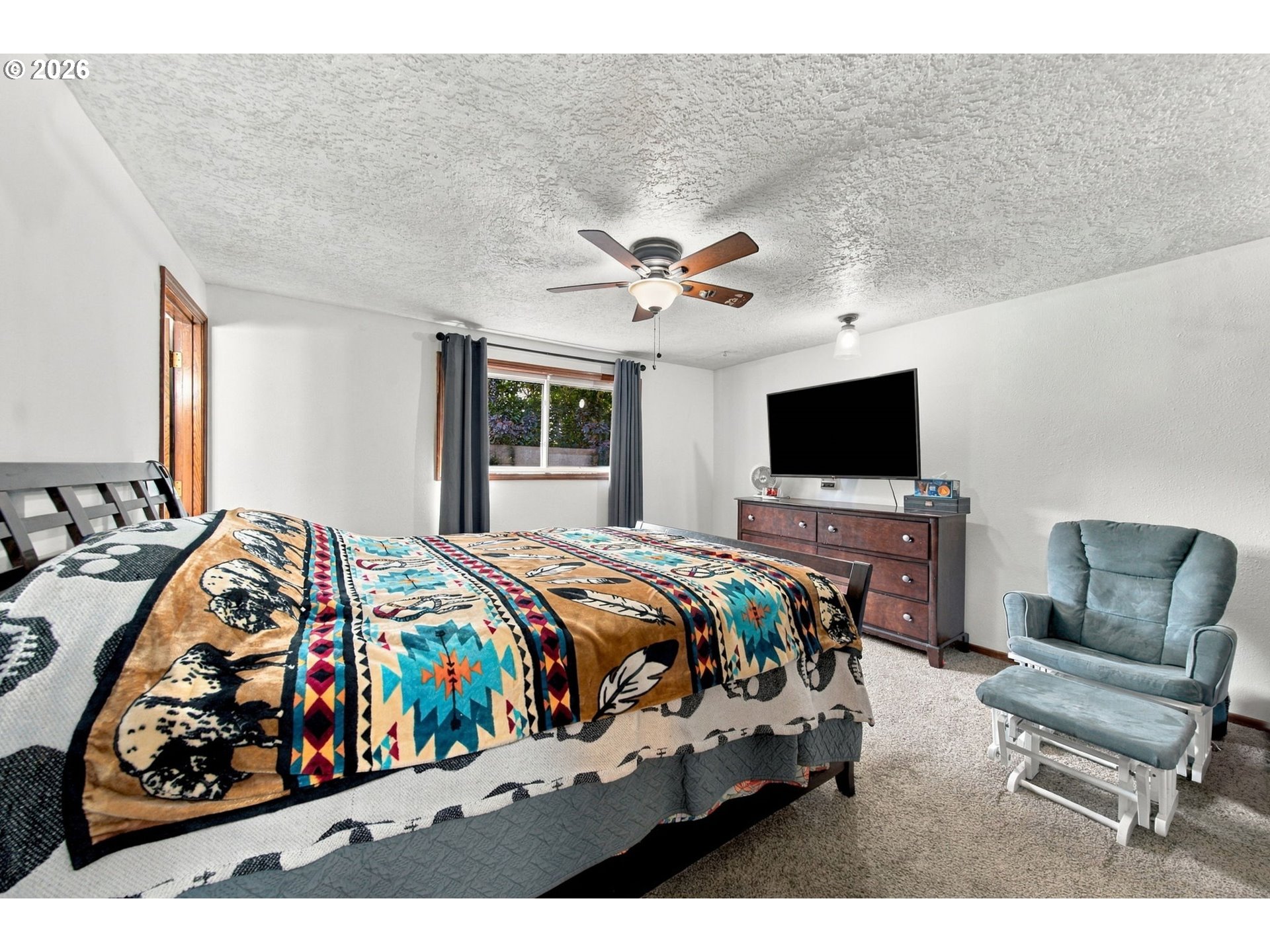 Furnished bedroom with a colorful patterned bedspread, ceiling fan, dresser, and blue-gray armchair.