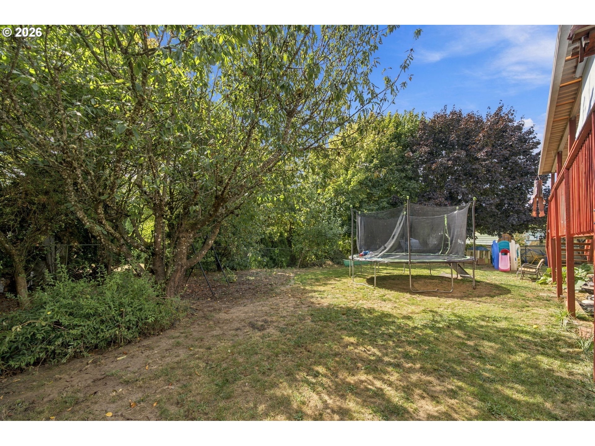 Backyard with mature shade trees, a trampoline, children’s play equipment, and a raised red deck.