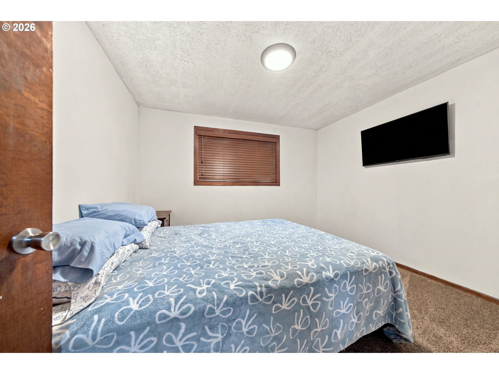 Bedroom with a blue patterned bedspread, wood blinds, and a wall-mounted television.