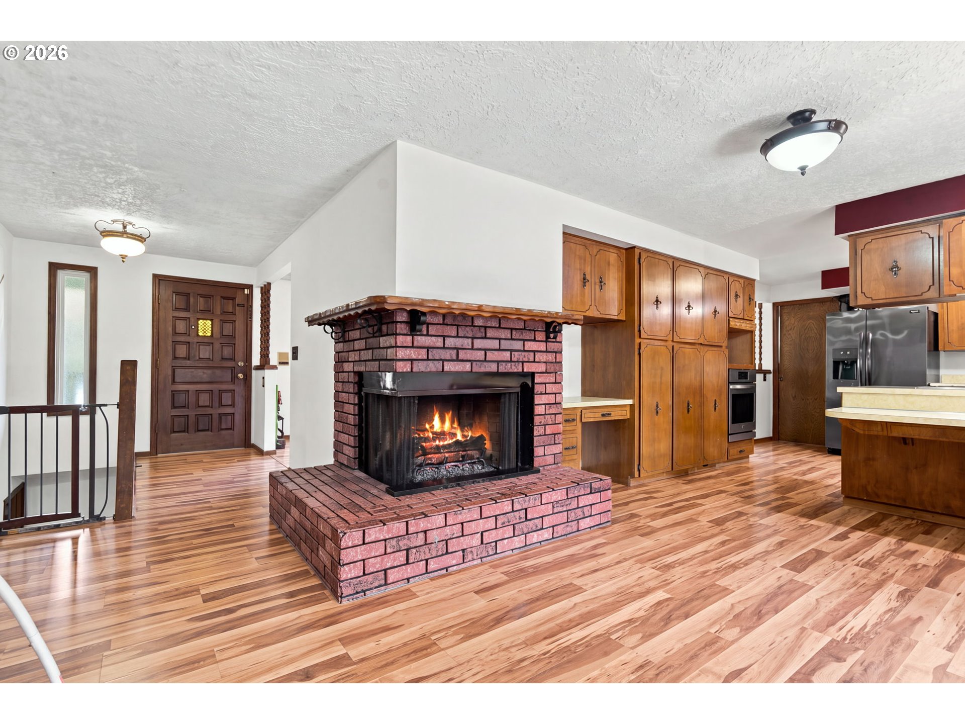 Open-plan living room and kitchen with a central brick fireplace, lit fire, wood cabinetry, and wood-look floors.