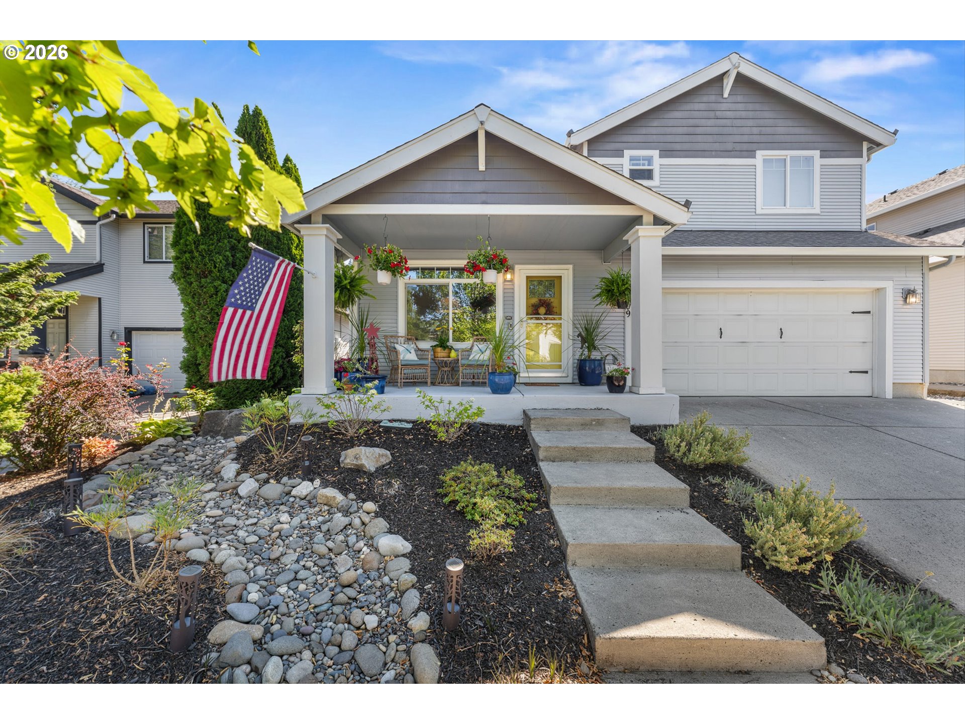 Gray two-story home with a covered, plant-filled porch, American flag, and landscaped rock garden.