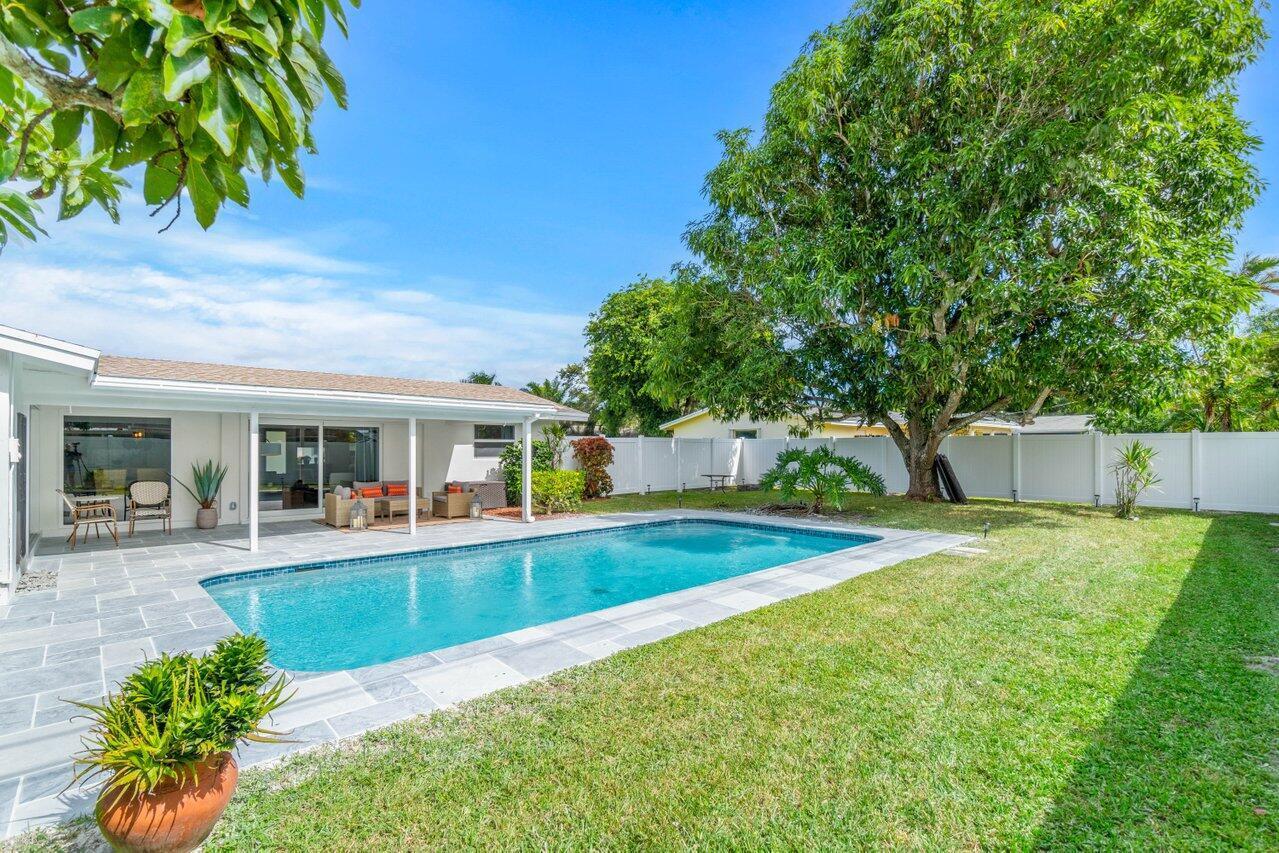 Backyard swimming pool beside a stone patio, lawn, white privacy fence, and mature shade tree.