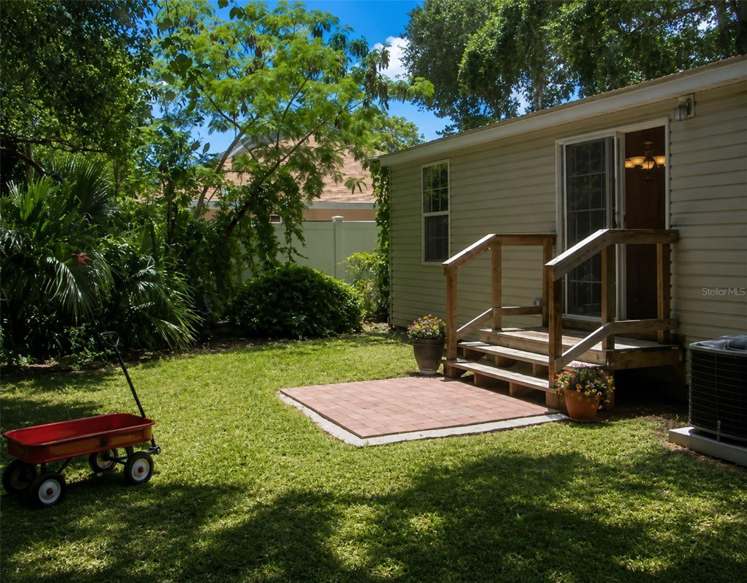 House backyard with a brick patio, wooden steps, lawn, mature trees, and a red wagon.