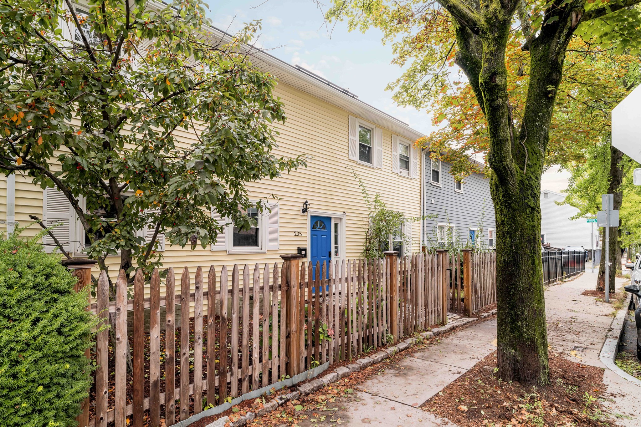 Yellow-sided home with a blue front door behind a wooden picket fence and mature street trees.