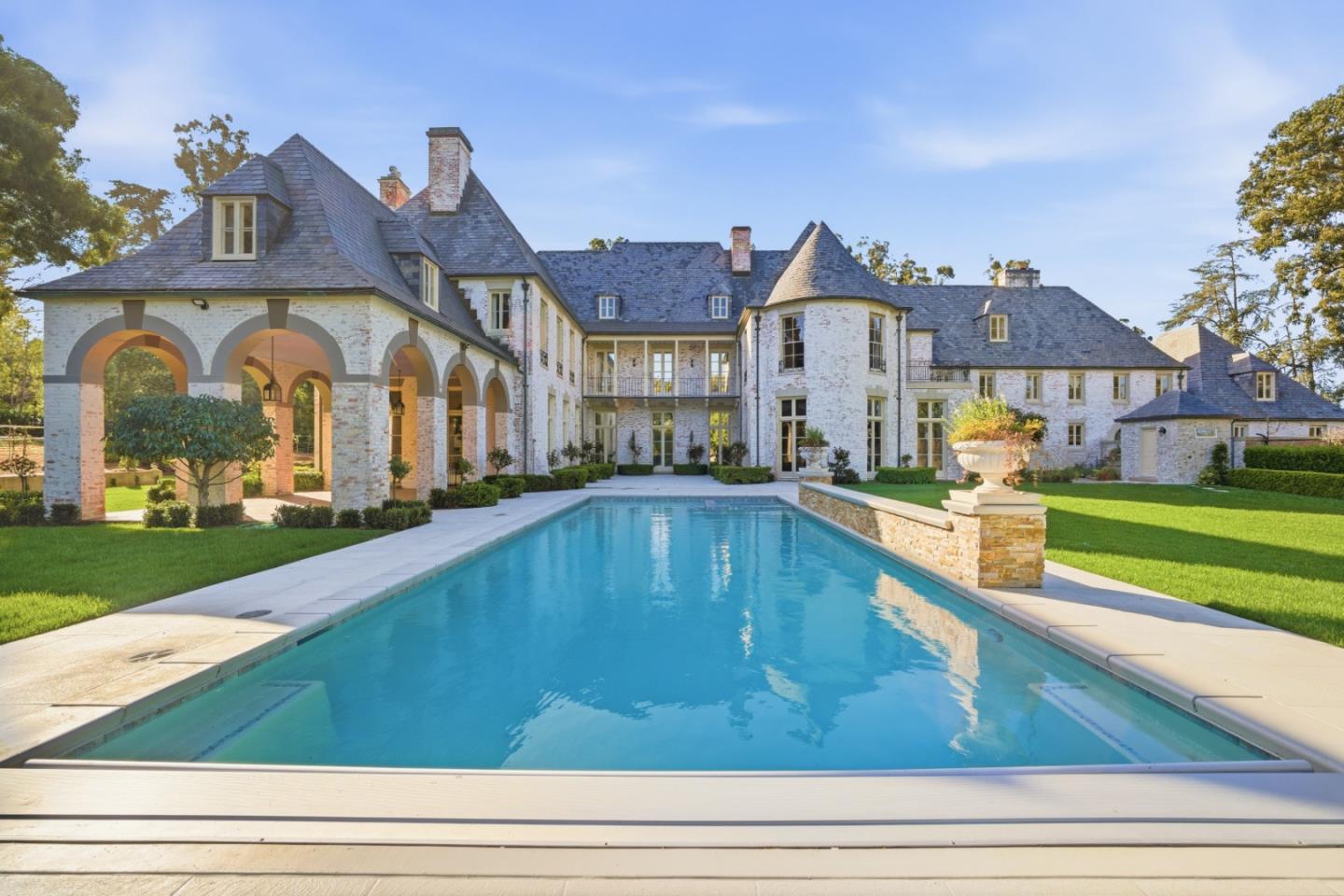Large white-brick estate with slate roofs and arched walkways behind a long swimming pool.