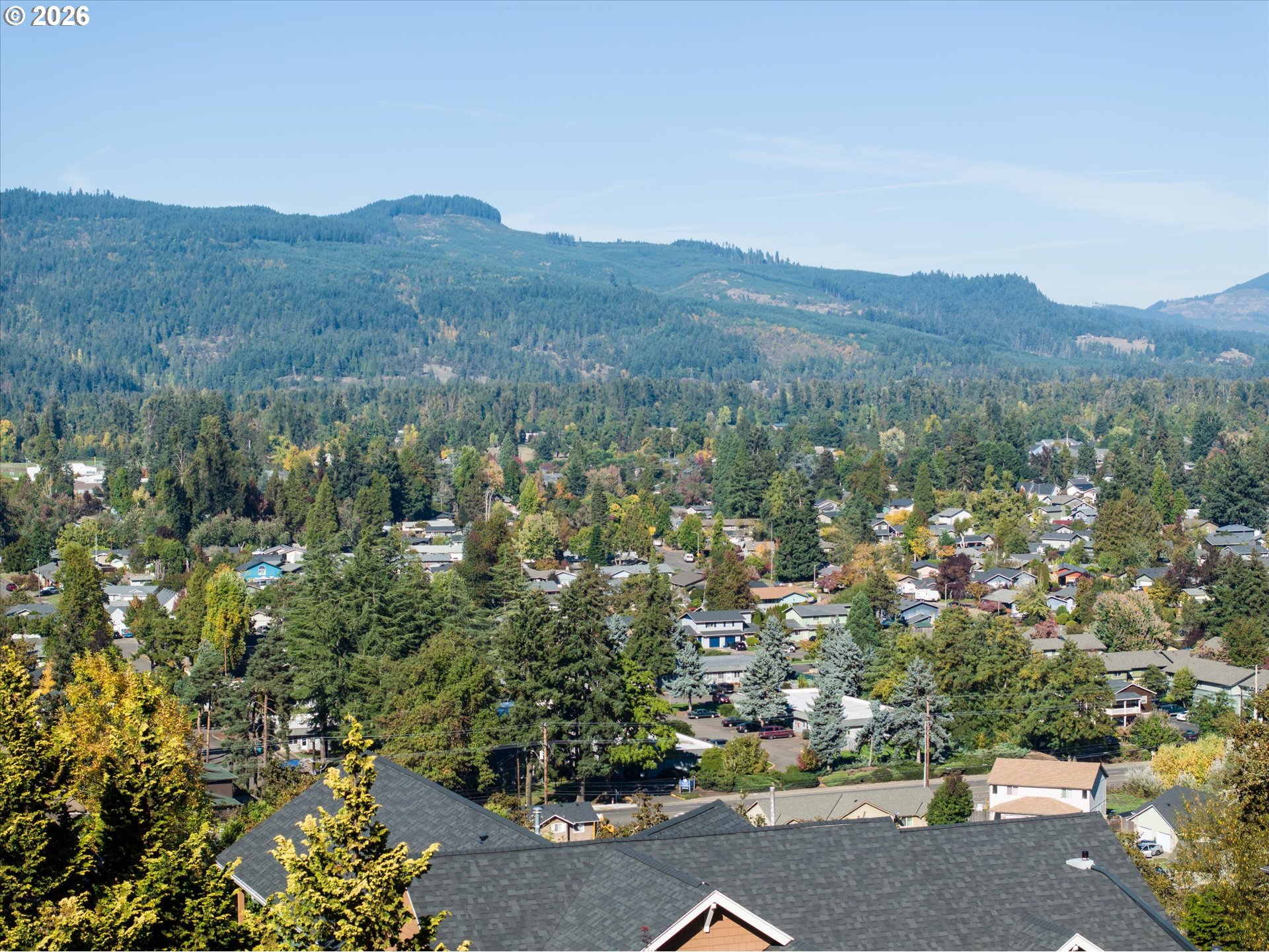 Residential neighborhood beneath forested mountains, viewed from above with autumn-colored trees.