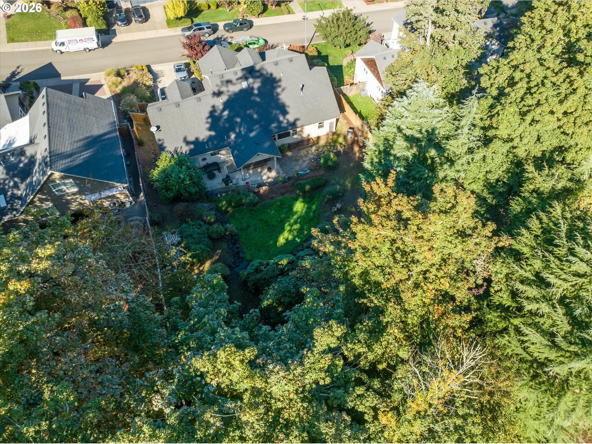 Aerial view of a house with a wooded backyard, lawn, patio, and narrow creek.