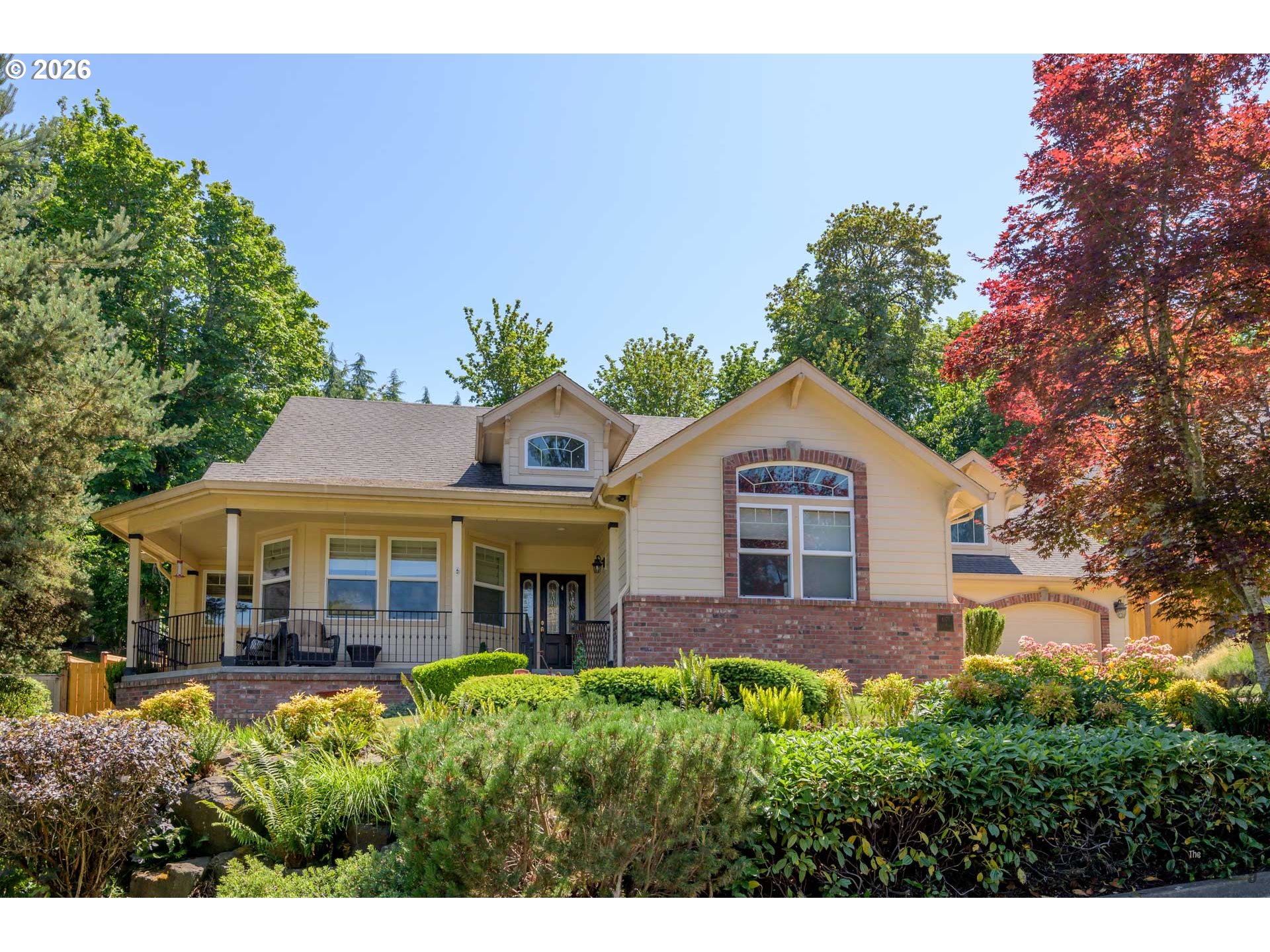 Beige house with brick accents, covered porch, and lush landscaping beneath a red-leaved tree.