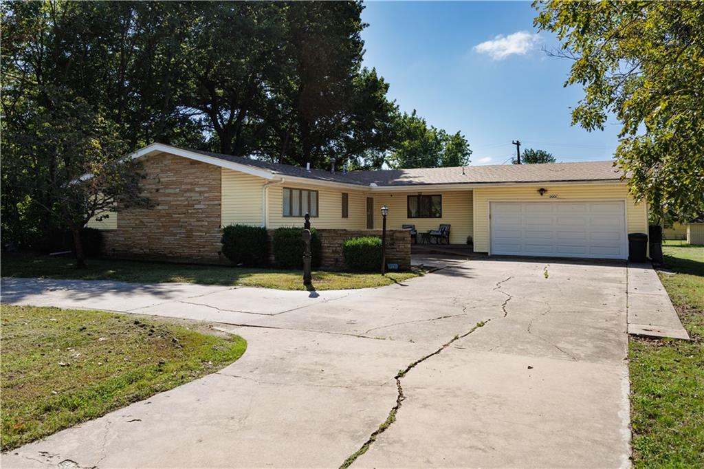 Single-story yellow ranch house with stone façade, attached garage, and wide cracked concrete driveway.