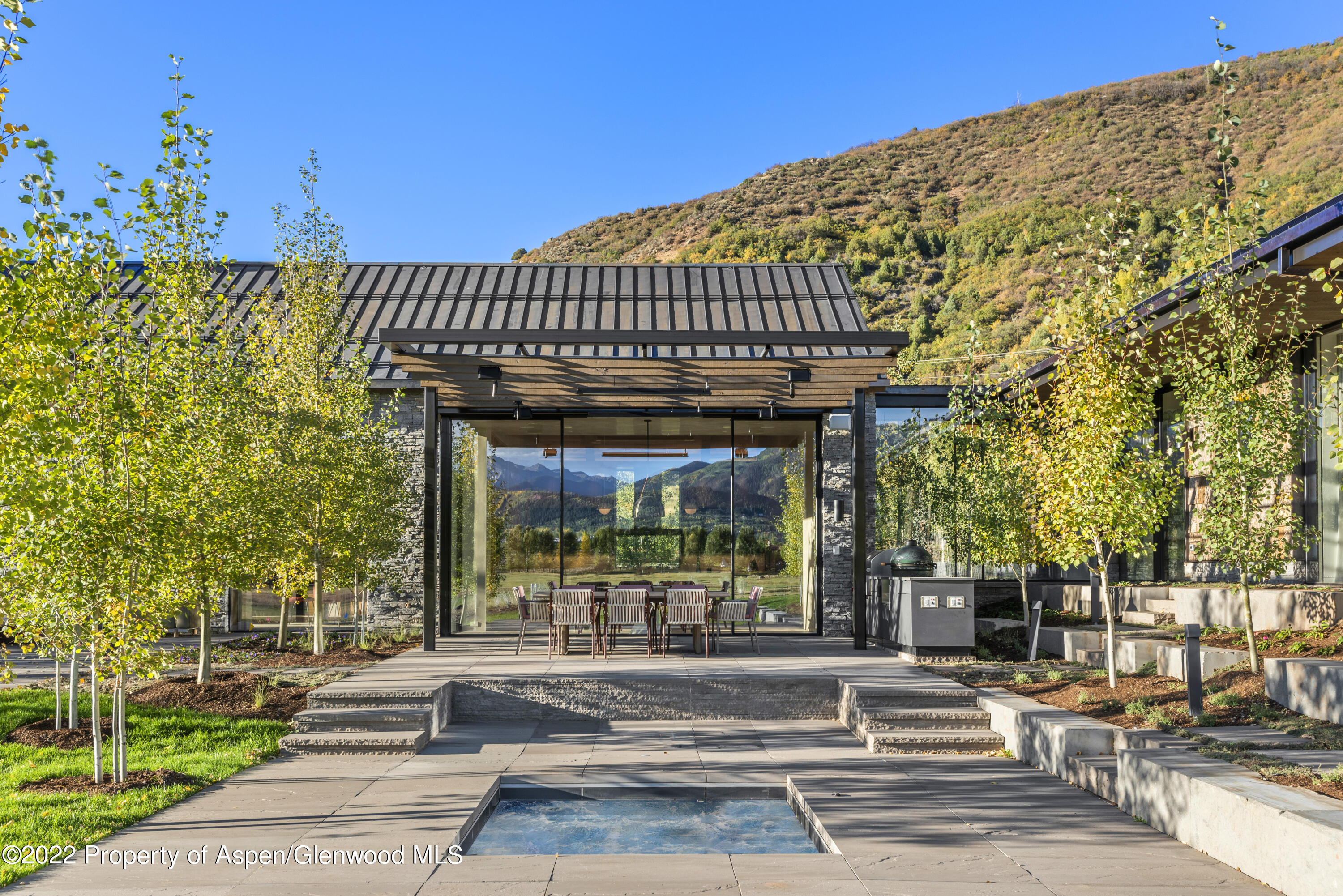 Modern glass-and-stone home with a patio dining area and reflecting pool, framed by autumn trees and mountain scenery.